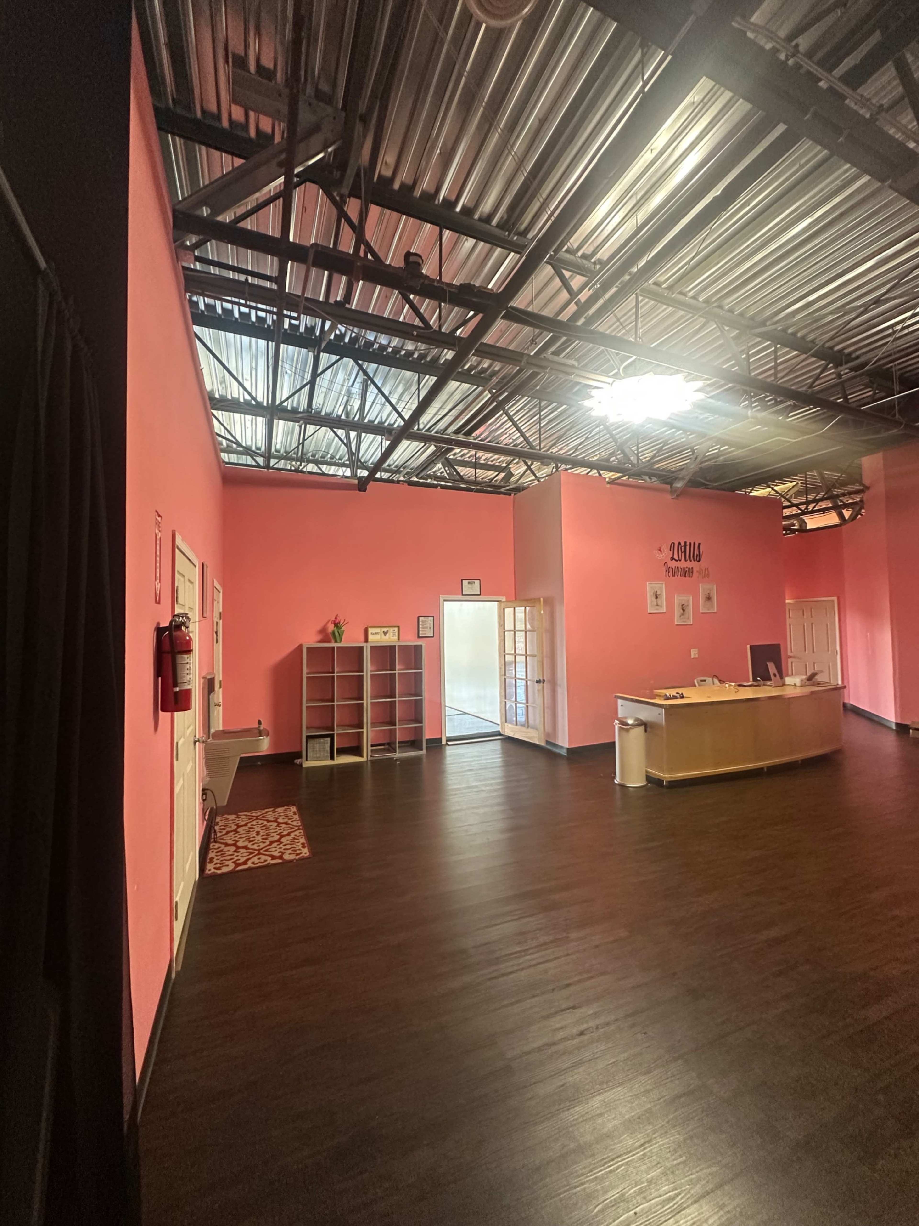 The image shows a spacious interior room with pink walls, dark wooden flooring, and a reception area at the back.