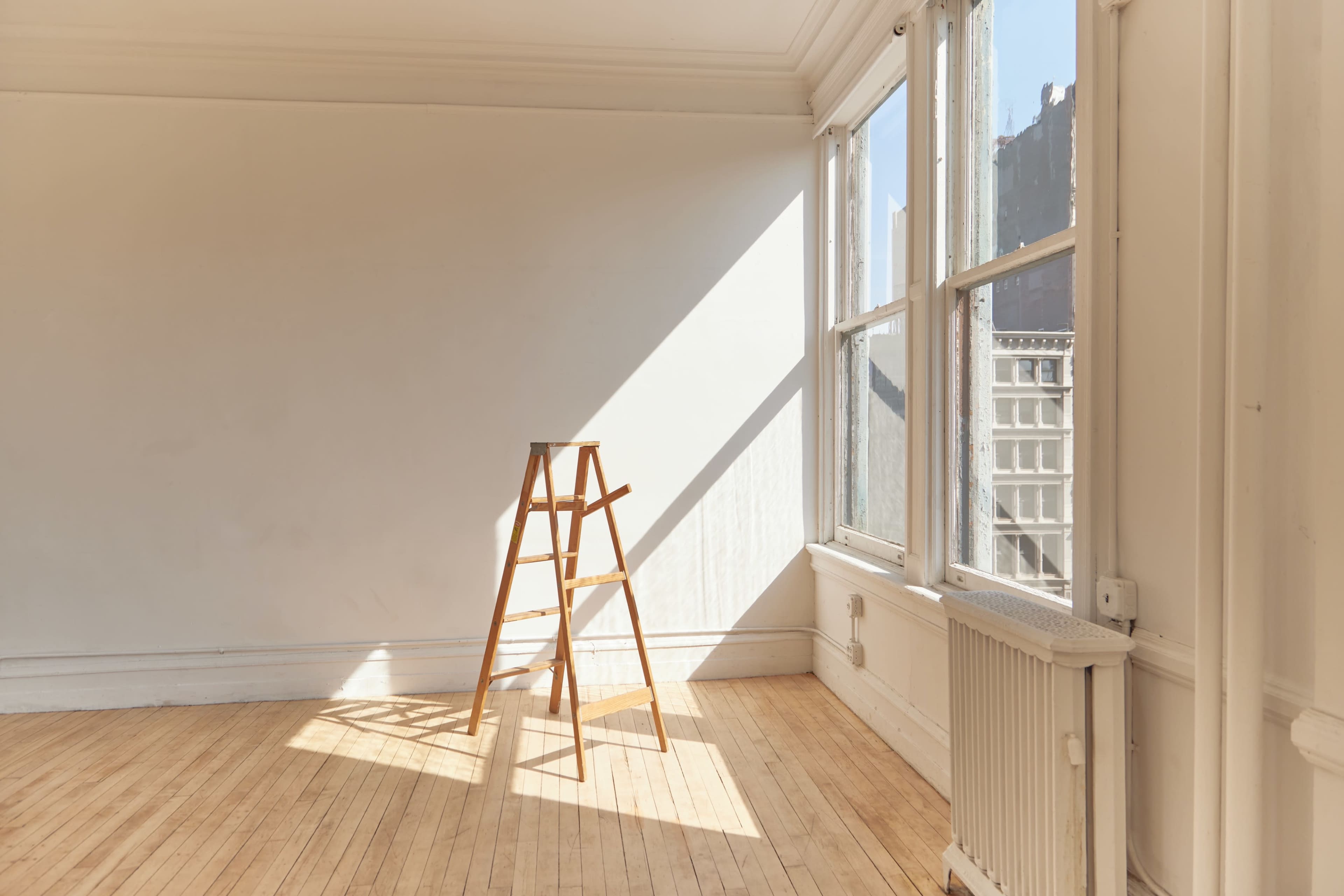 A wooden ladder stands in an empty room with large windows allowing sunlight to stream in.