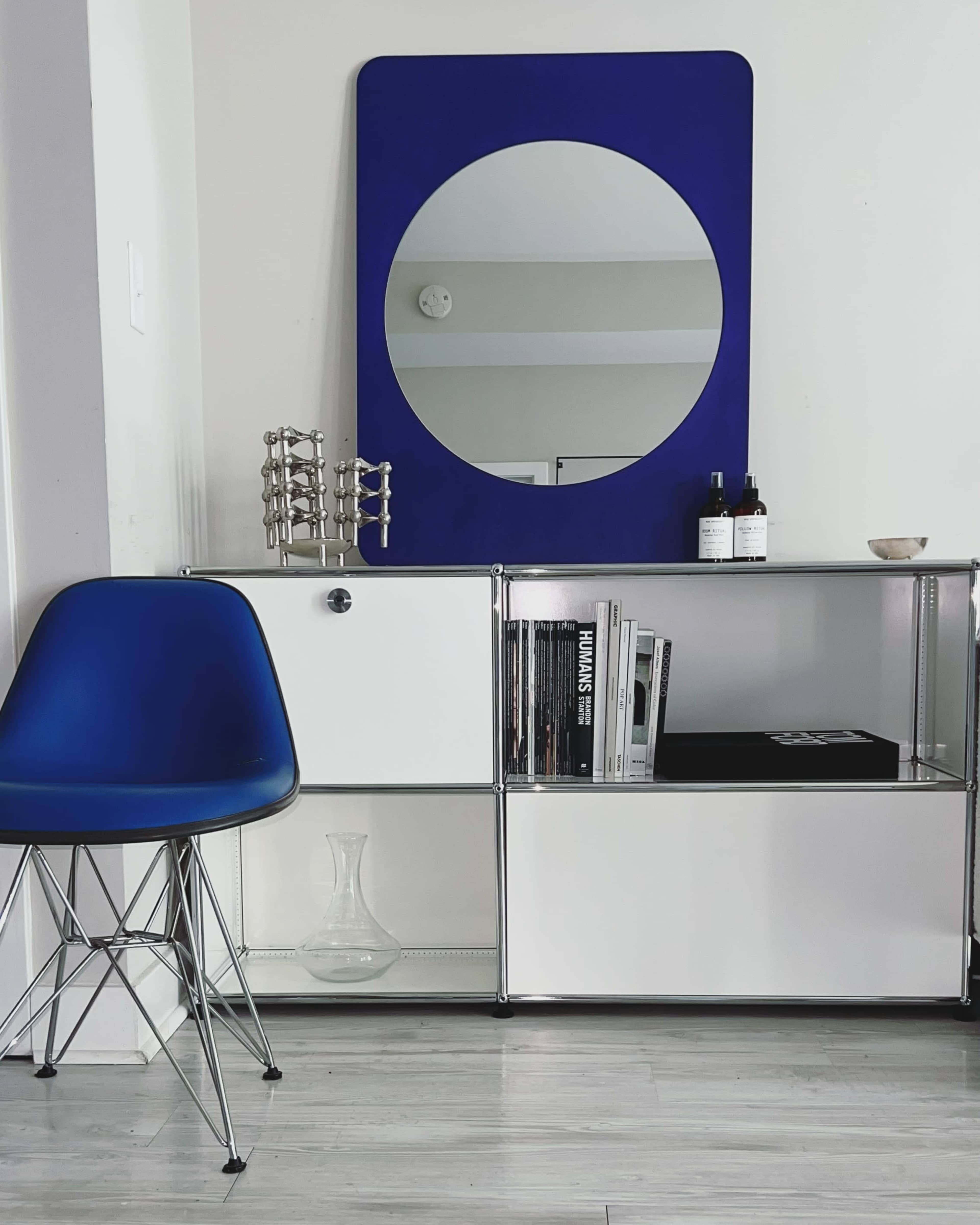 A modern interior scene featuring a blue chair next to a white and chrome shelving unit with a circular mirror above it.