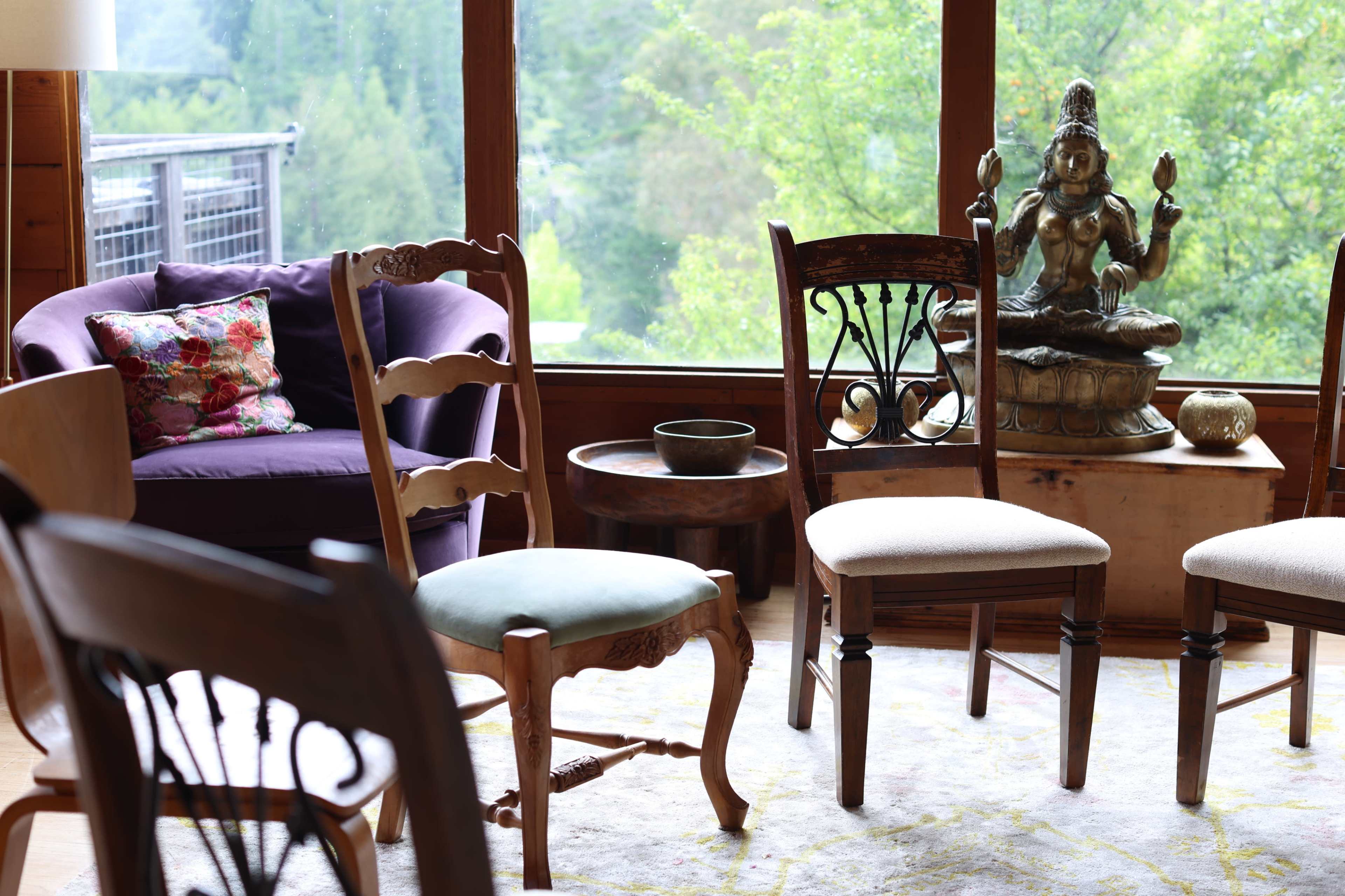 The image depicts a cozy interior with various styled chairs, a purple couch, and a statue of a seated figure surrounded by large windows showcasing greenery outside.