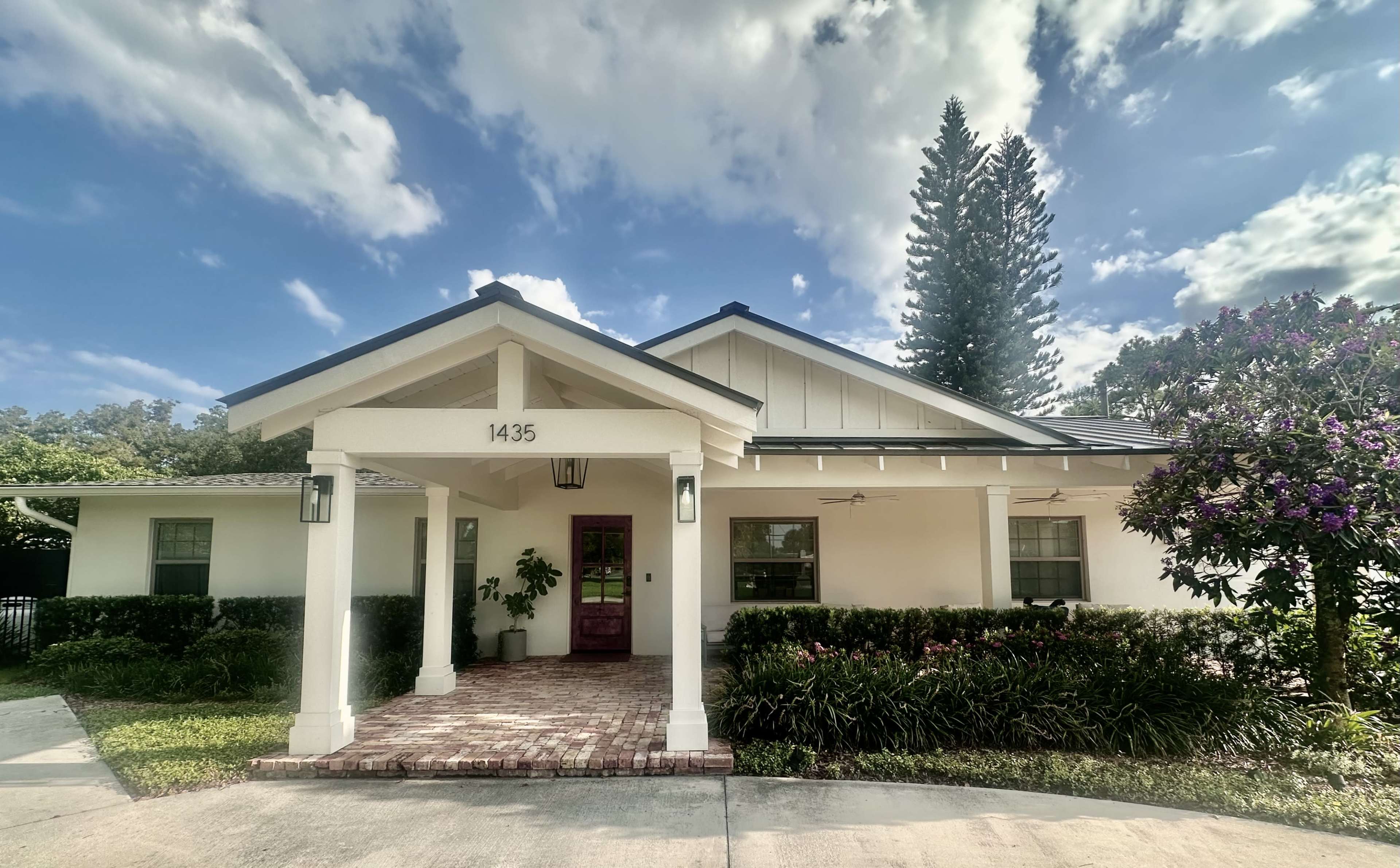 A single-story house with a sloped roof and a front porch is set among greenery and trees.