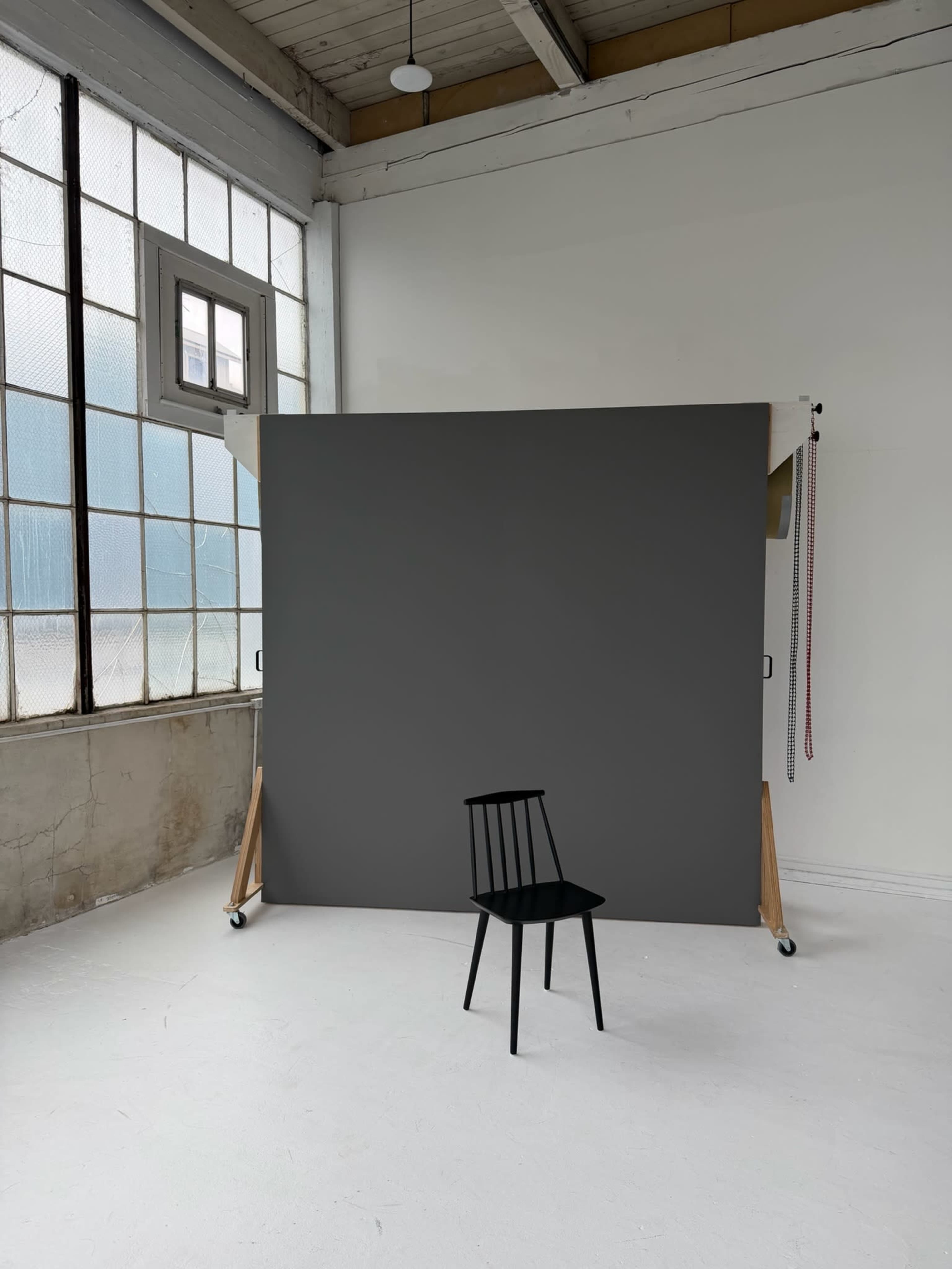 A black chair is placed in front of a gray backdrop within a bright, industrial-style room featuring large windows.