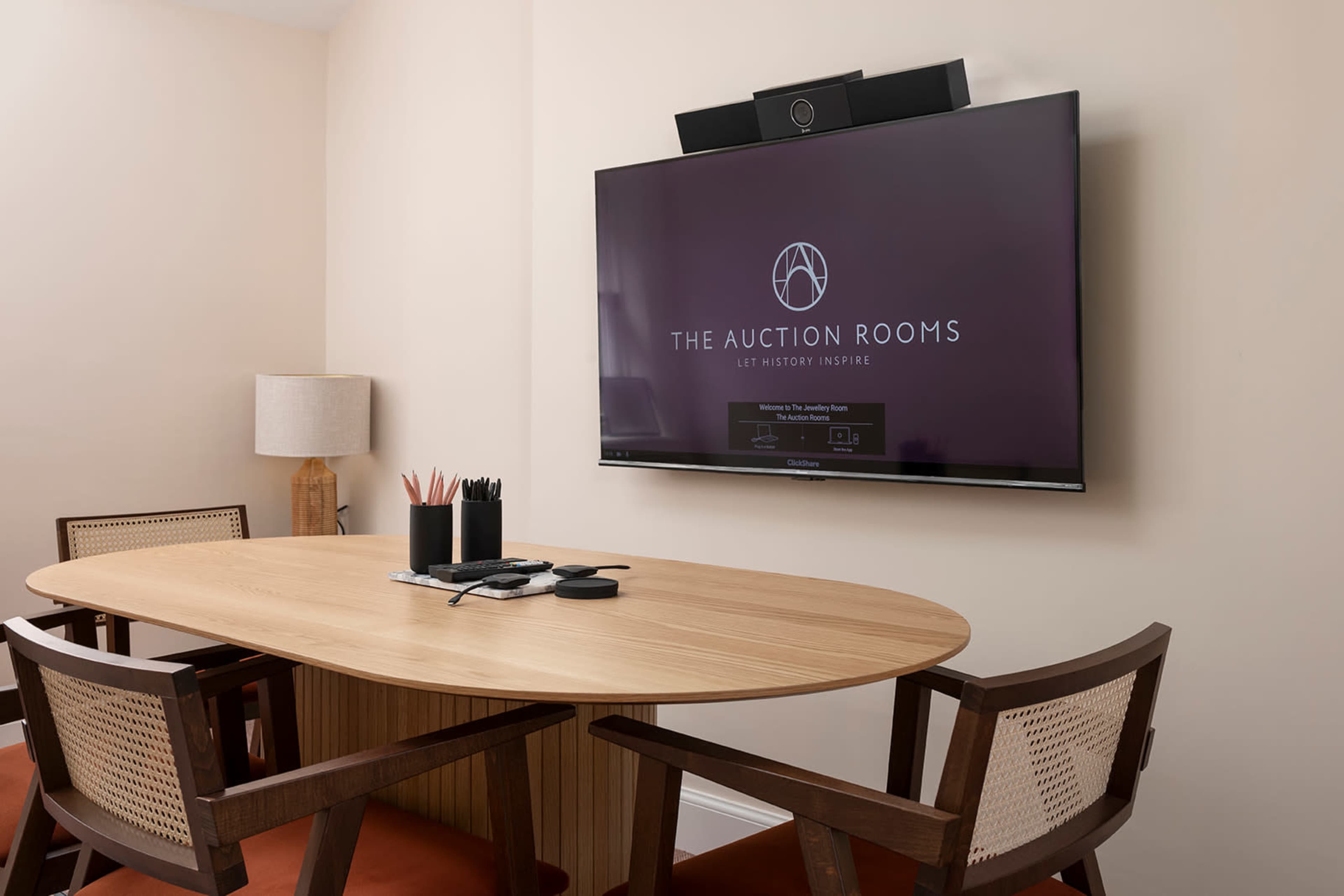 A circular wooden conference table is set with office supplies, and a large television display is mounted on the wall, featuring the logo of "The Auction Rooms."