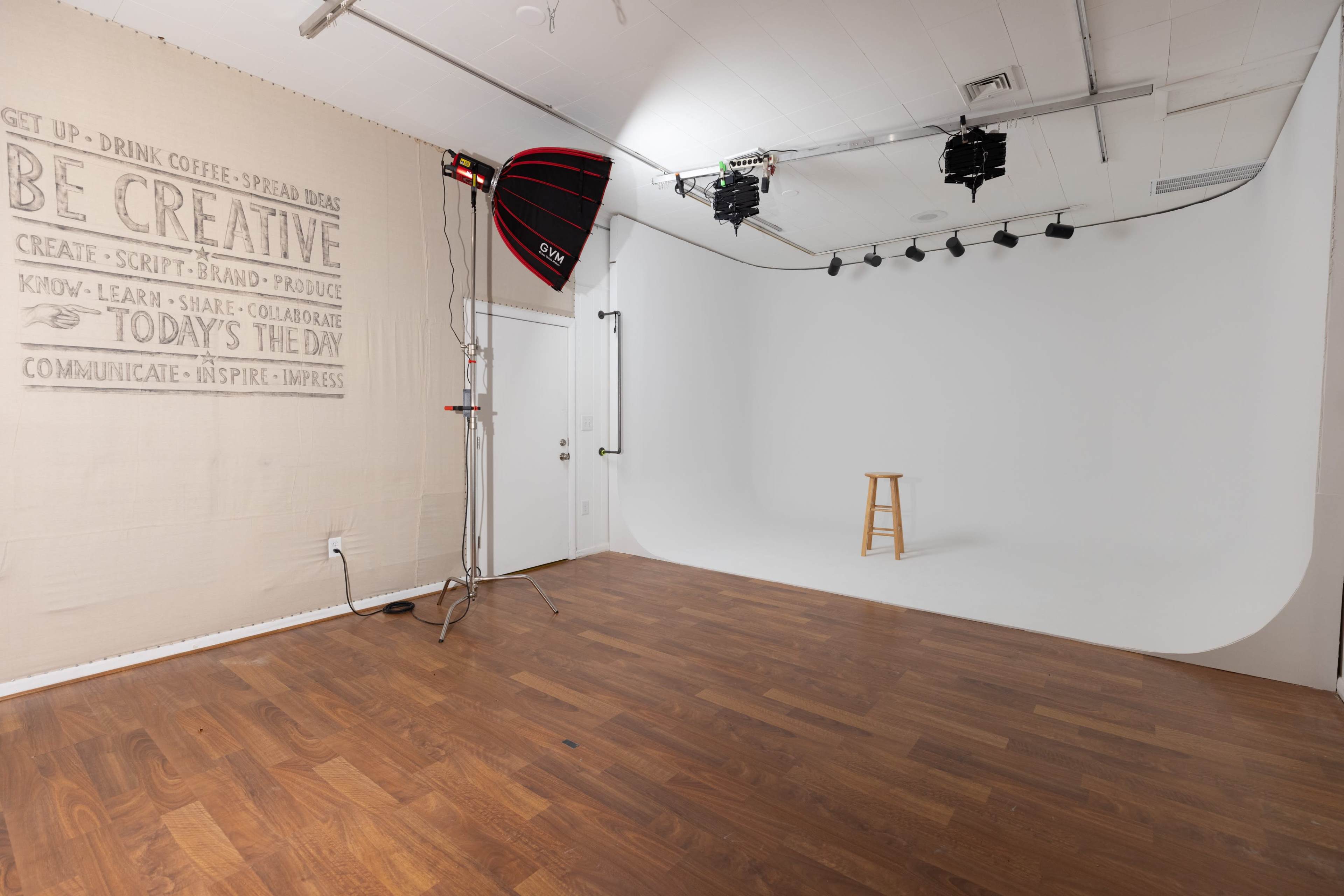 A photography studio features a white backdrop, wooden flooring, and a stool in the center, with lighting equipment set up along one side.