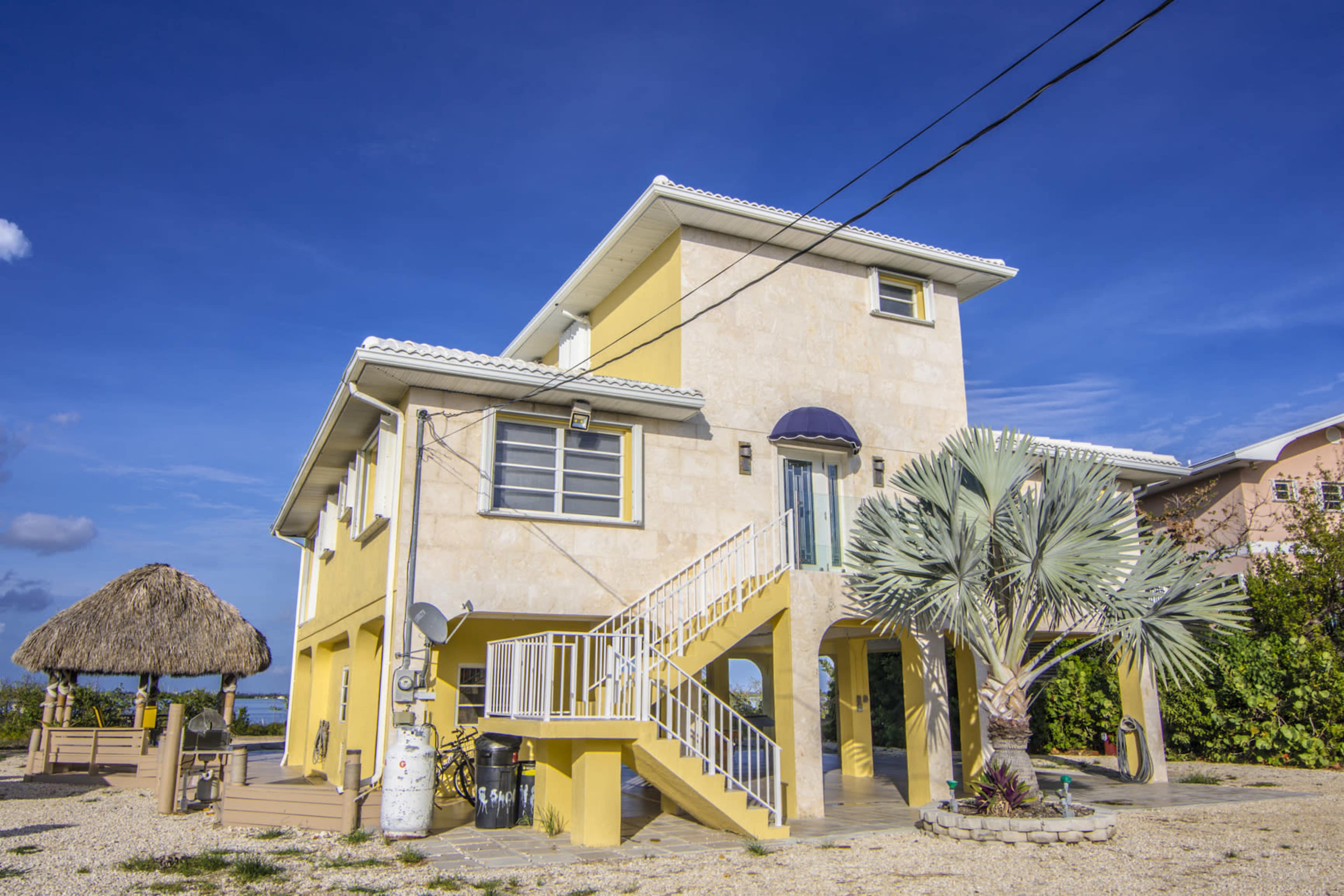 The image shows a two-story yellow house with a porch and a thatched-roof gazebo nearby, set against a clear blue sky.