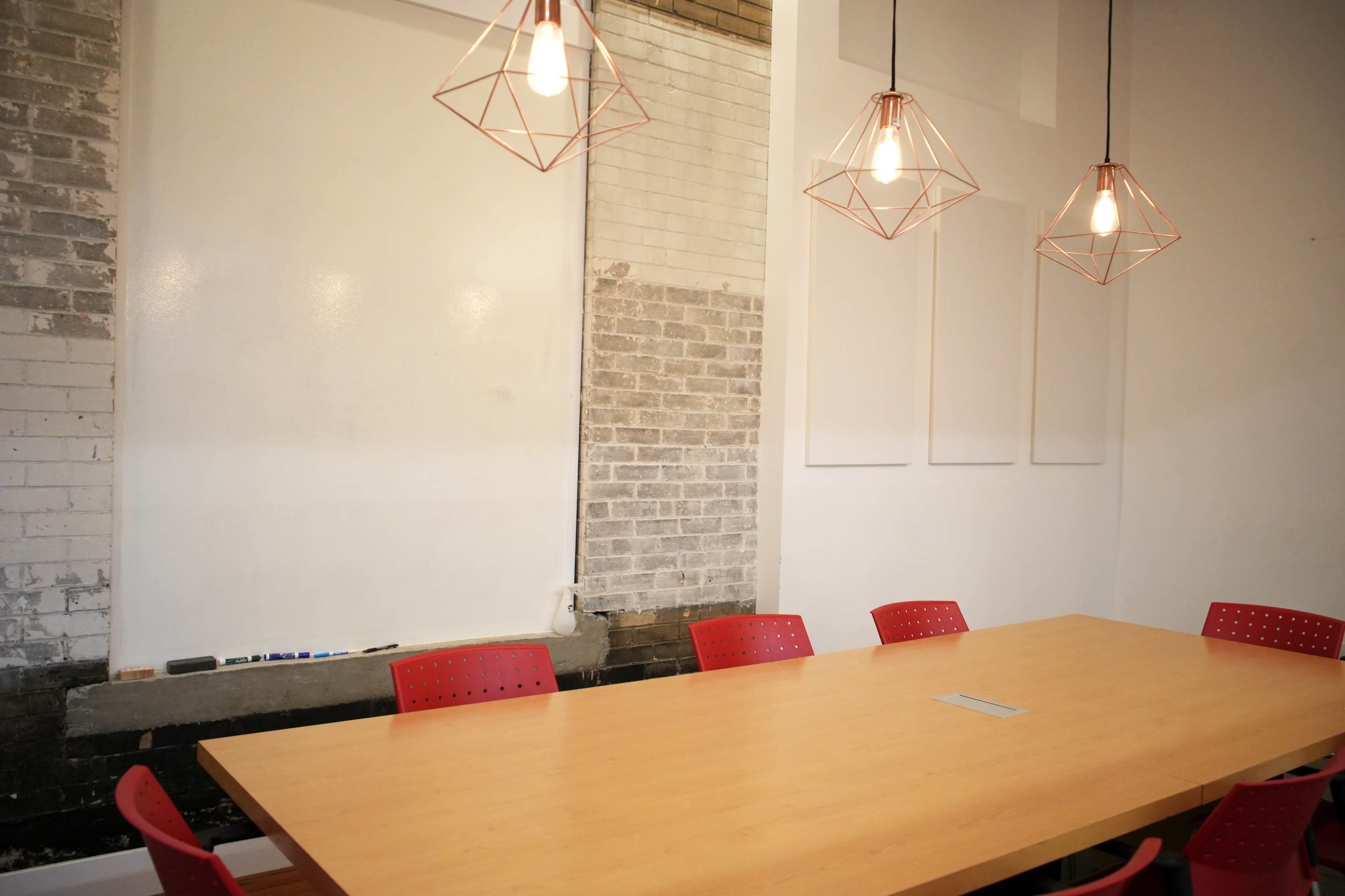 A spacious meeting room features a wooden table surrounded by red chairs, with three hanging light fixtures and a blank whiteboard on the wall.