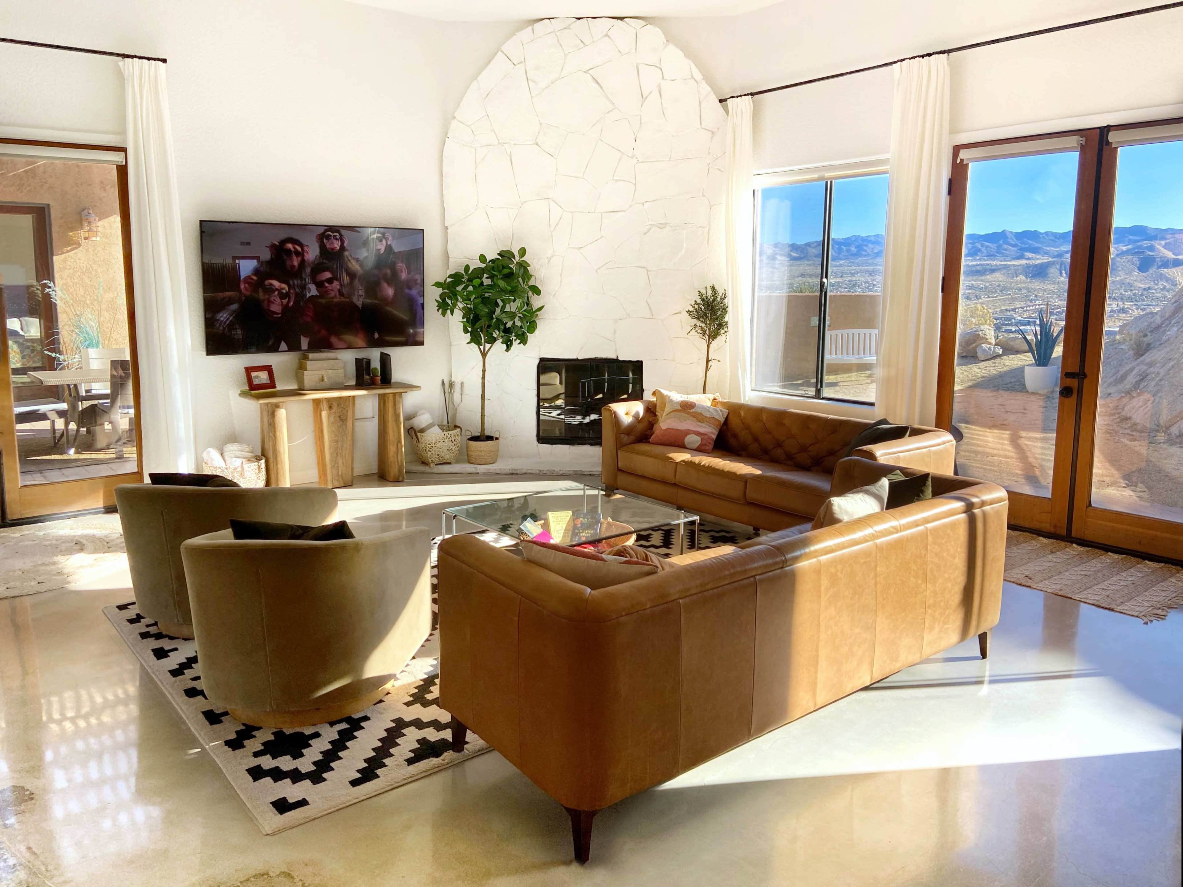 A modern living room with a large window revealing a desert landscape, featuring brown leather sofas, a glass coffee table, and a television mounted on the wall.