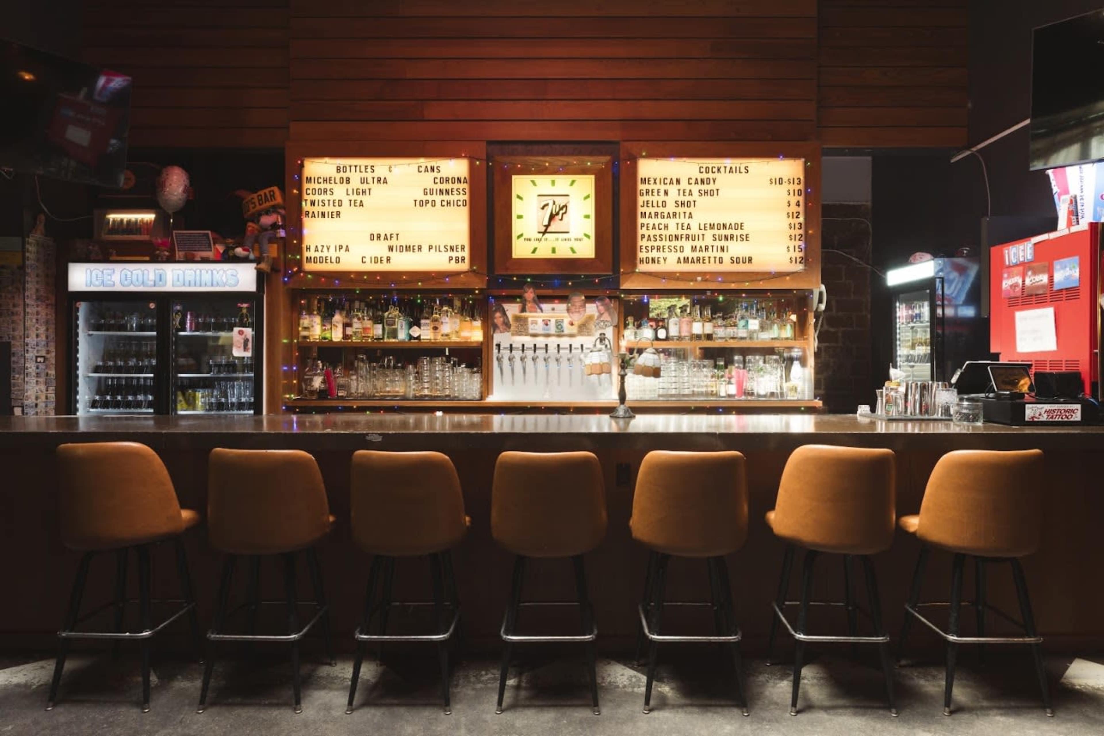 A bar with orange stools, illuminated signage displaying drink options, and shelves lined with glassware is set against a wooden wall backdrop.