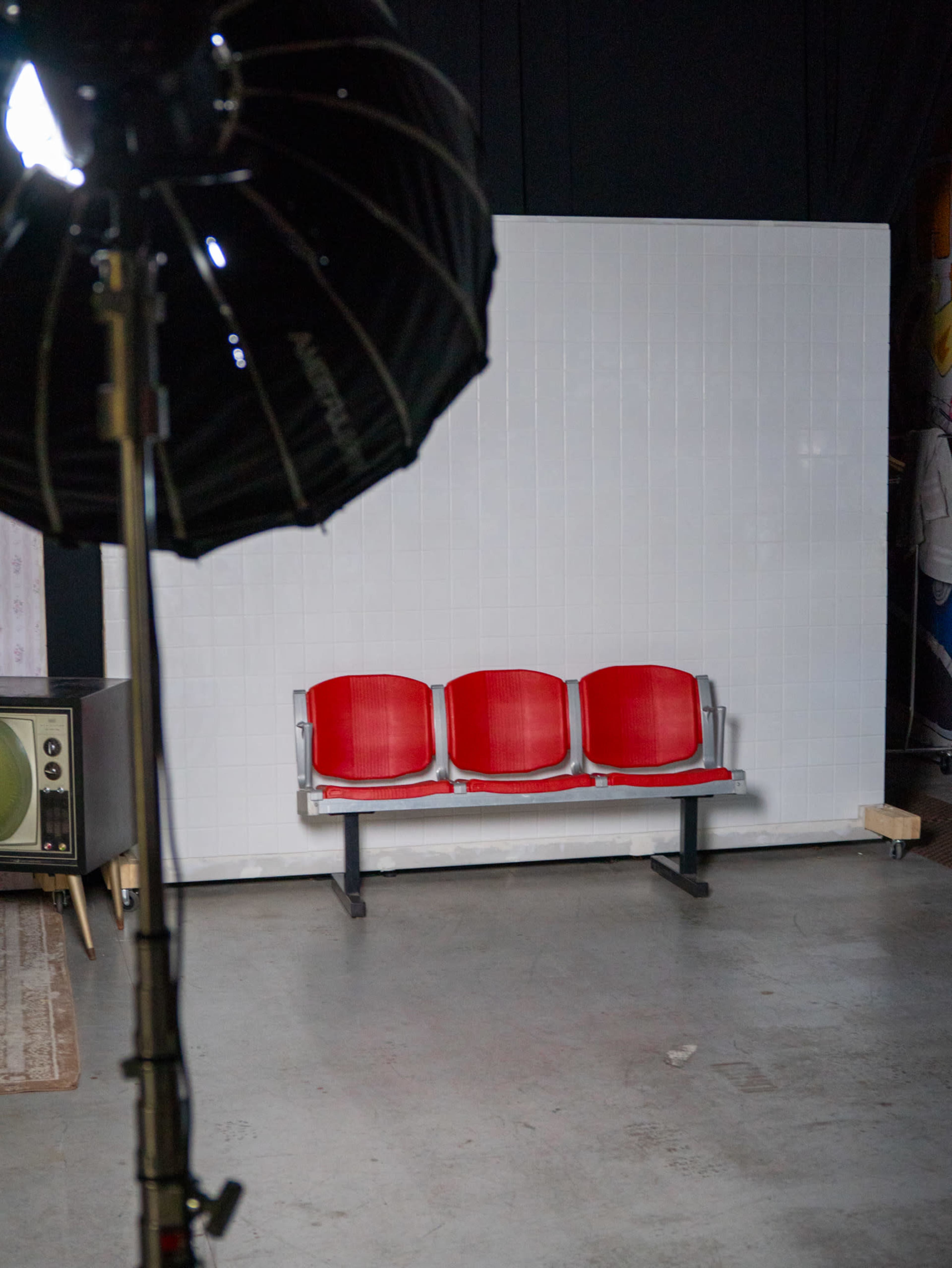 The image shows a three-seat red bench facing a white backdrop in an indoor setting, with a studio light in the foreground.