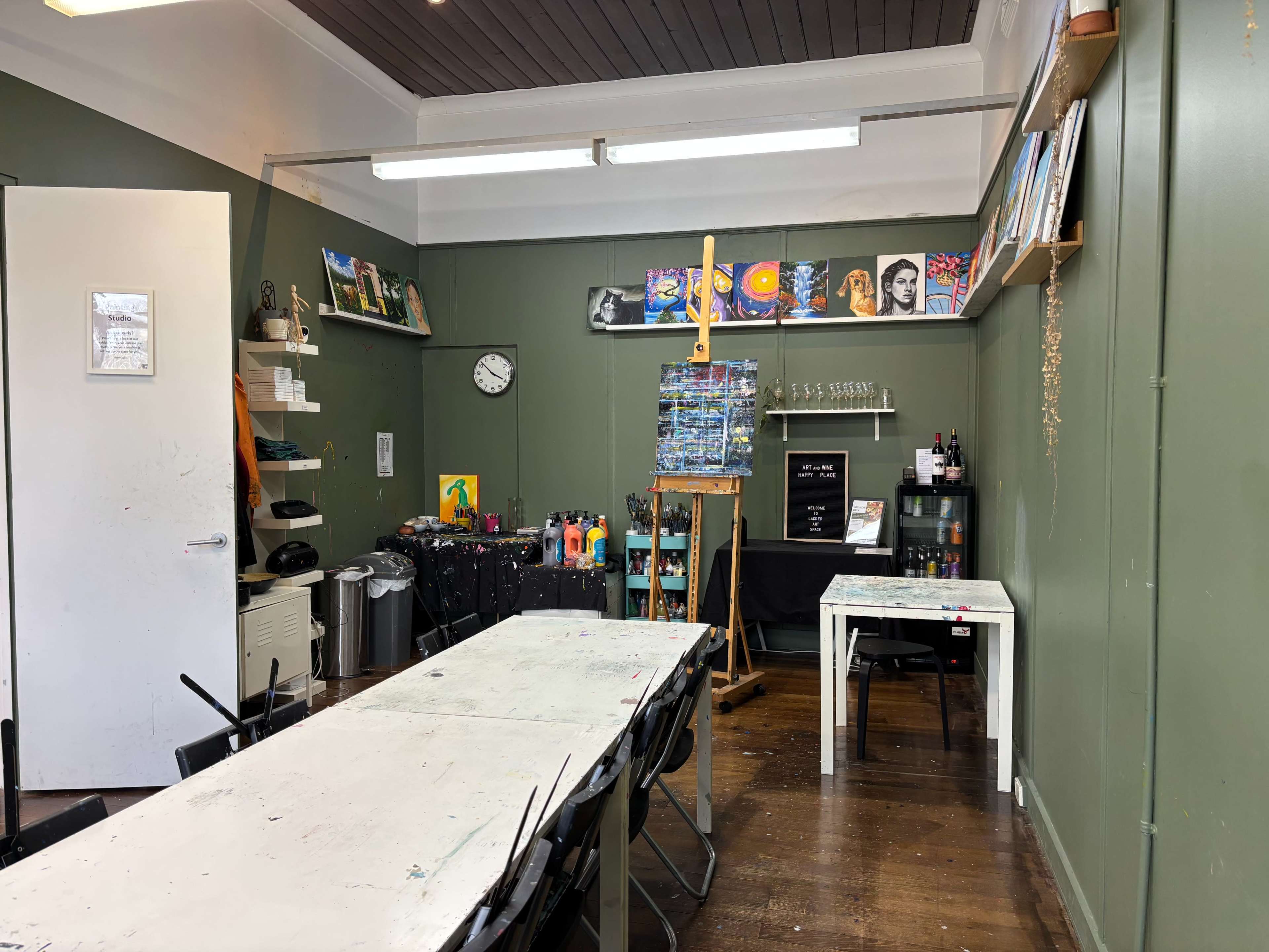 The image shows an art studio with green walls, a large table surrounded by chairs, an easel, art supplies, and a clock on the wall.