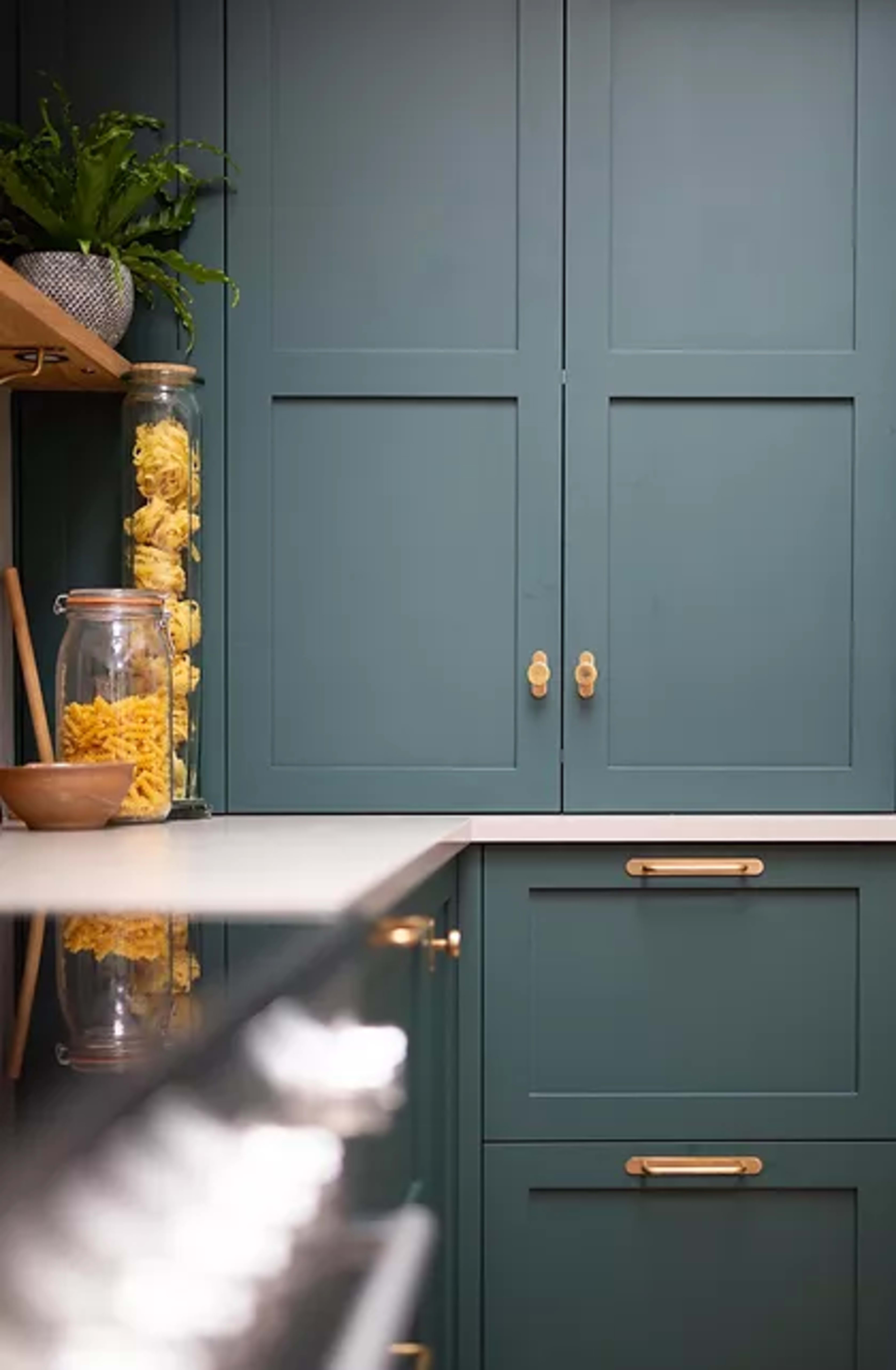 A kitchen scene features teal cabinetry with brass hardware, a glass jar filled with pasta, and a dark countertop reflecting the surroundings.