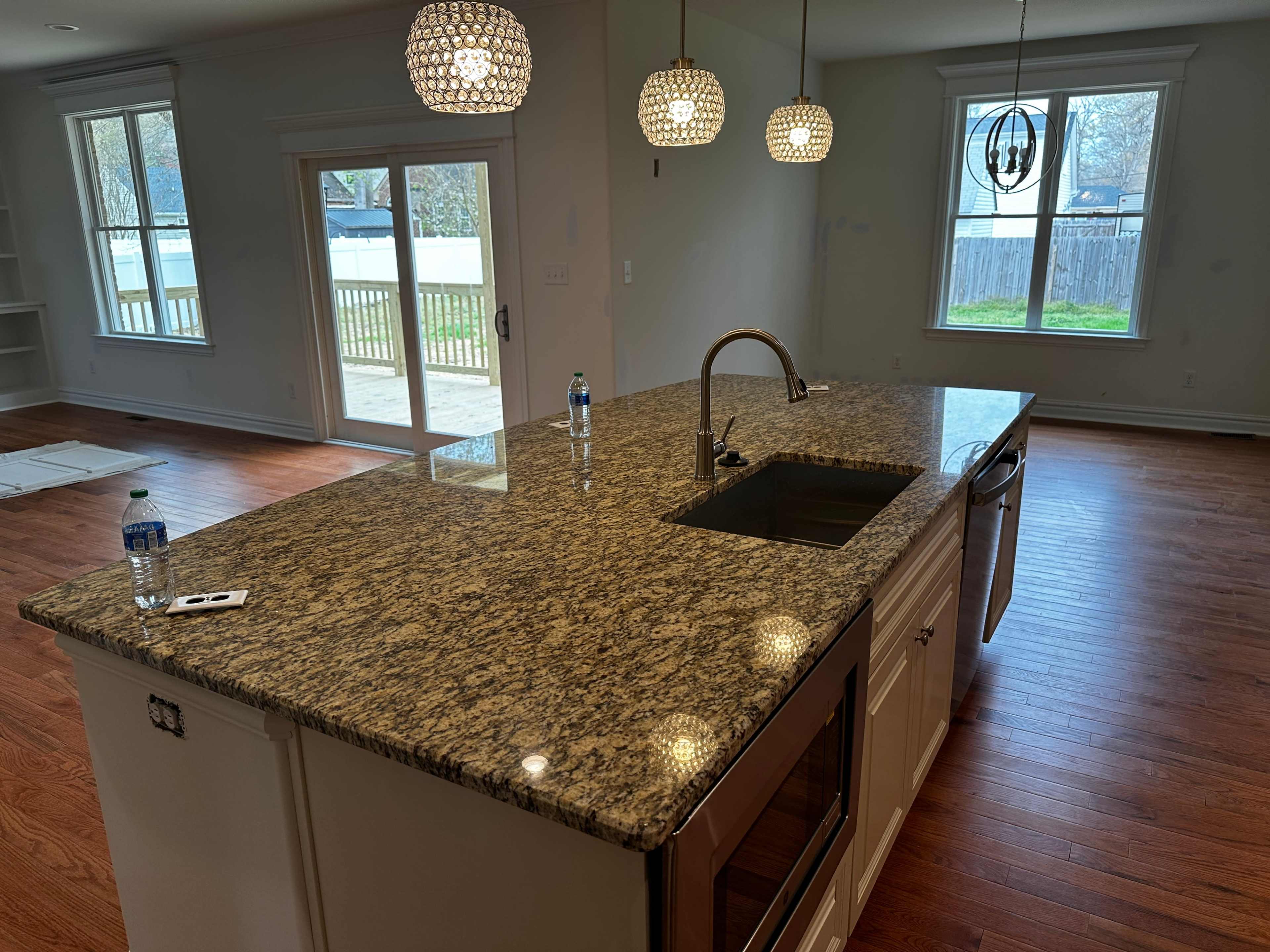 A kitchen with a granite countertop island featuring a sink, pendant lights overhead, and windows that overlook a backyard.