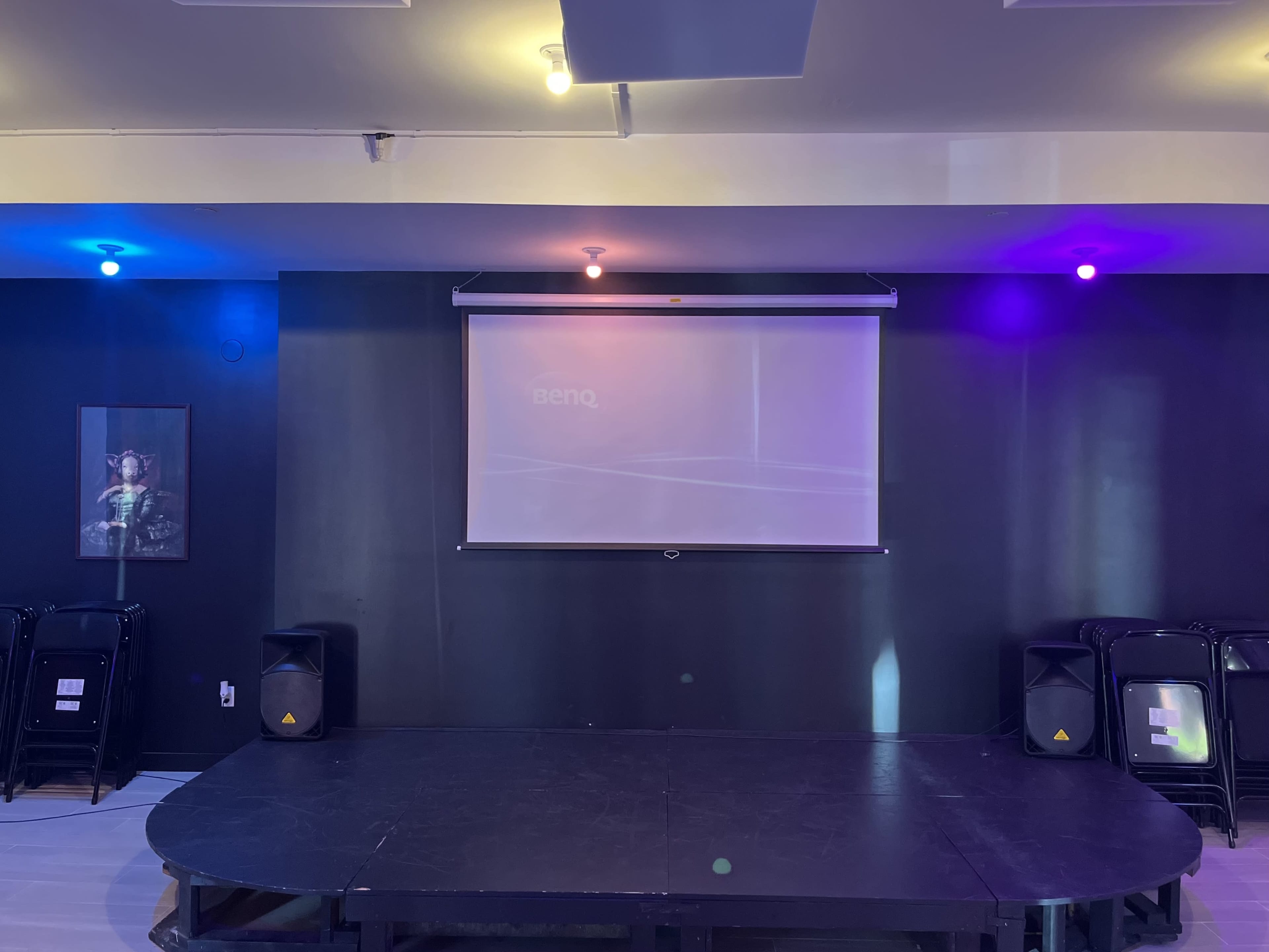 A stage area with a retractable projection screen and lighting in purple and blue hues, flanked by black folding chairs.