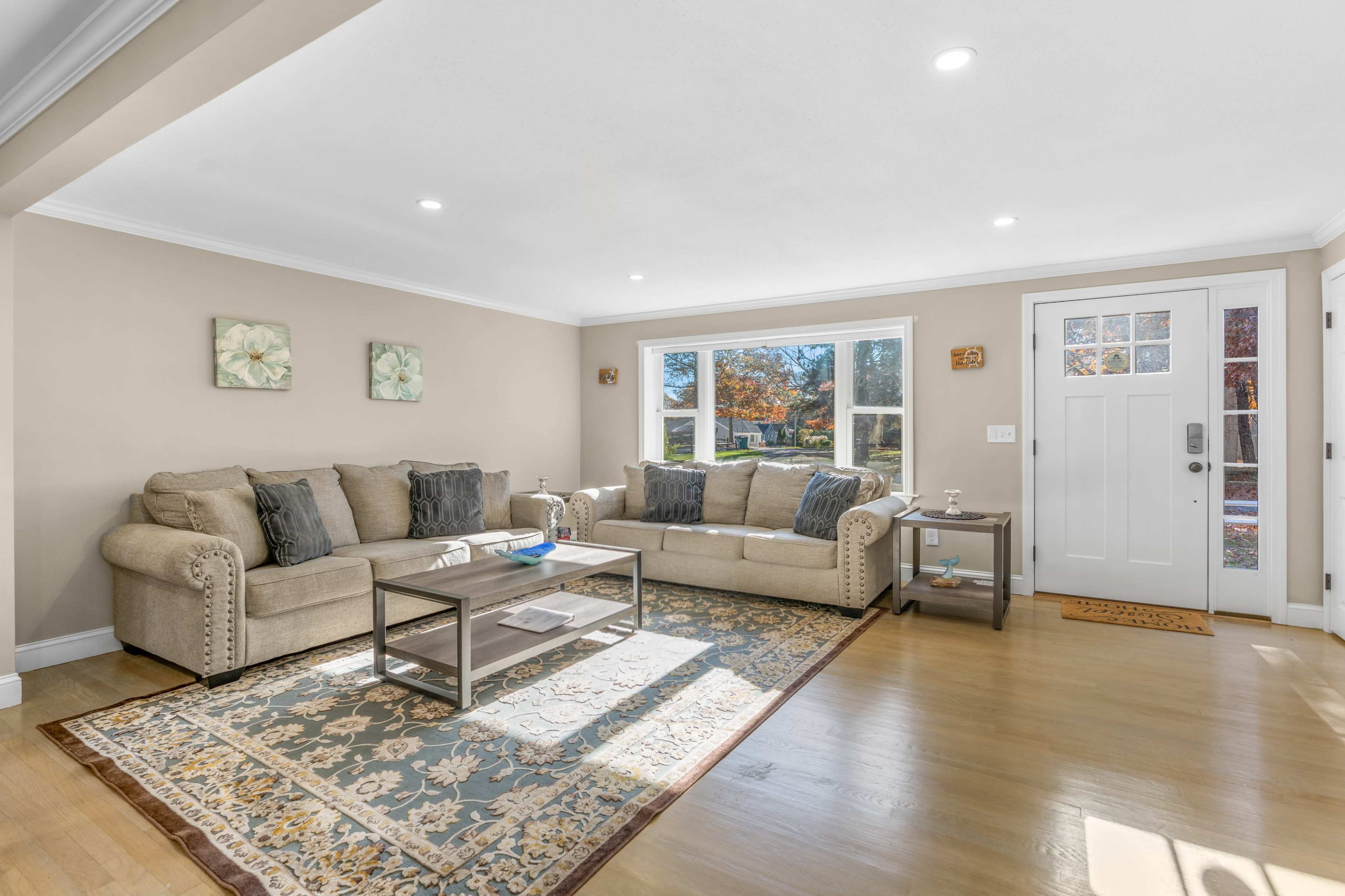 A spacious living room features a beige sectional sofa, a decorative area rug, a wooden coffee table, and large windows letting in natural light.