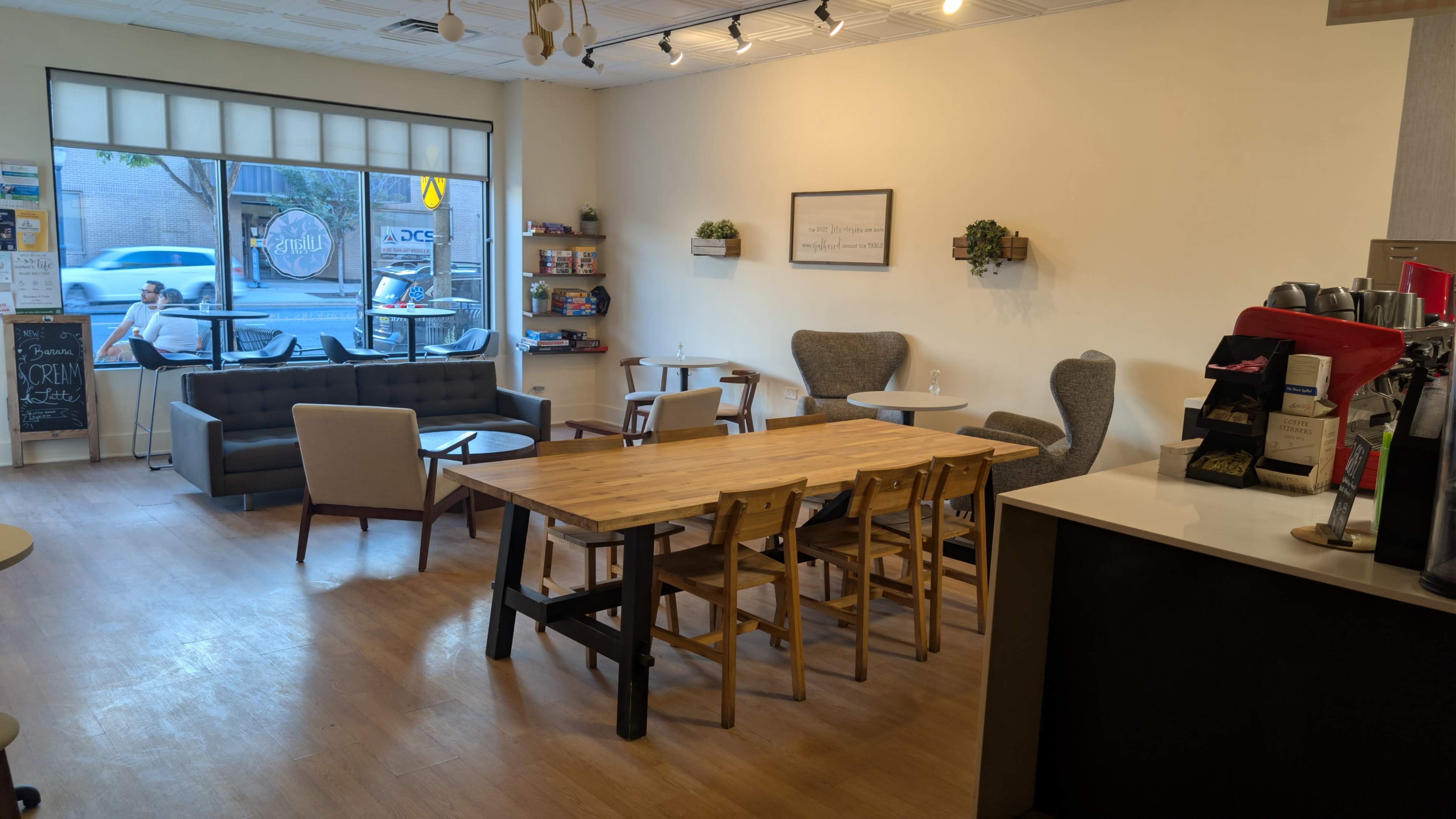 The image shows a cozy cafe interior featuring a wooden table surrounded by chairs, a grey sofa, and various seating options with natural light coming through large front windows.