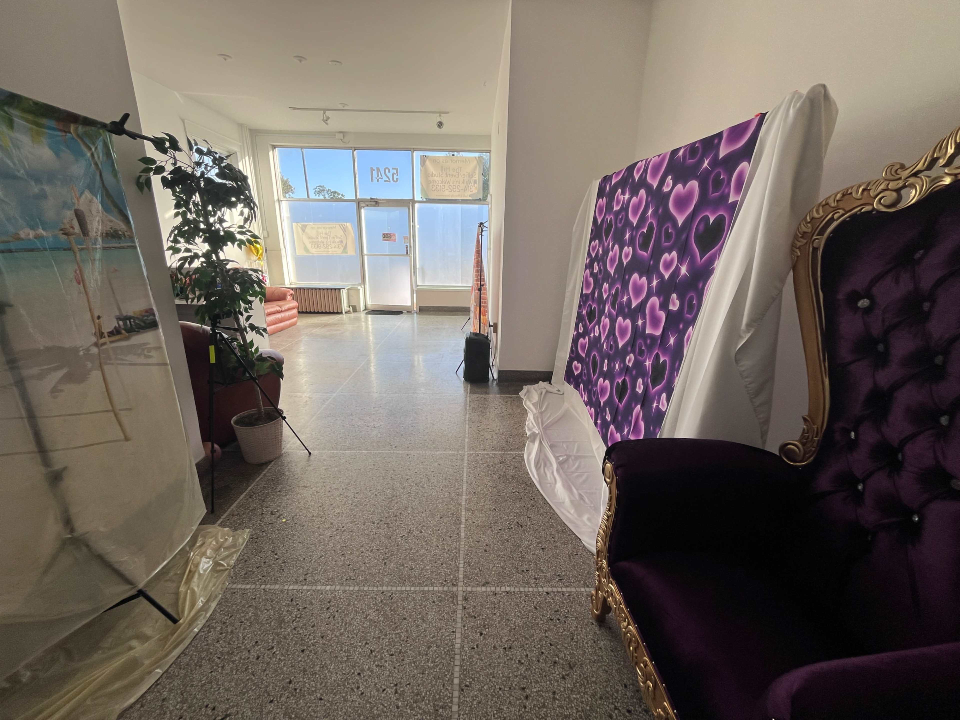 The image shows a bright hallway with a decorative purple chair, a large purple and pink heart-patterned backdrop, and a beach-themed backdrop on the left side.