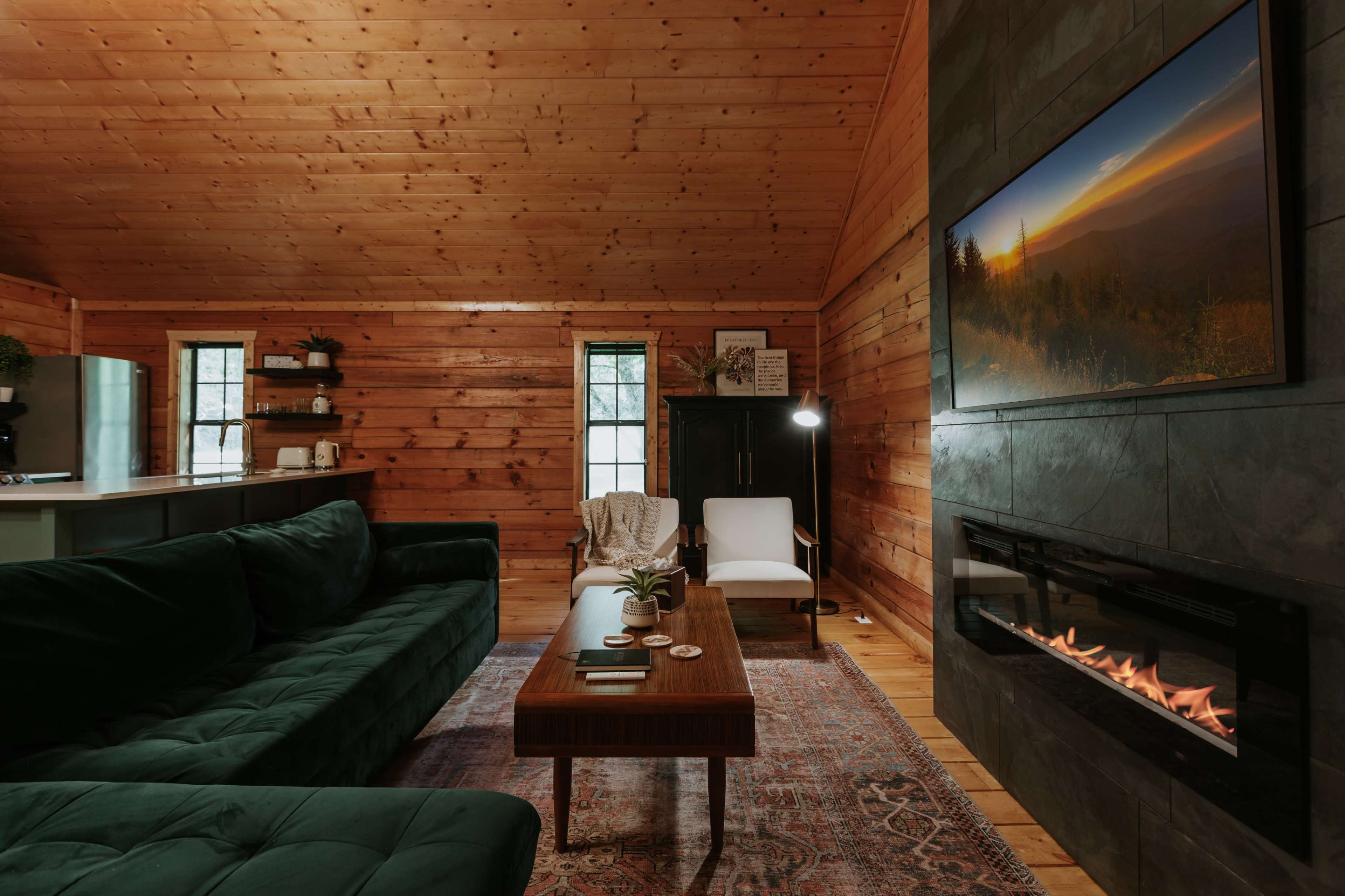 A cozy cabin living room featuring a green velvet sofa, a wooden coffee table, and a modern fireplace beneath a television displaying a scenic landscape.