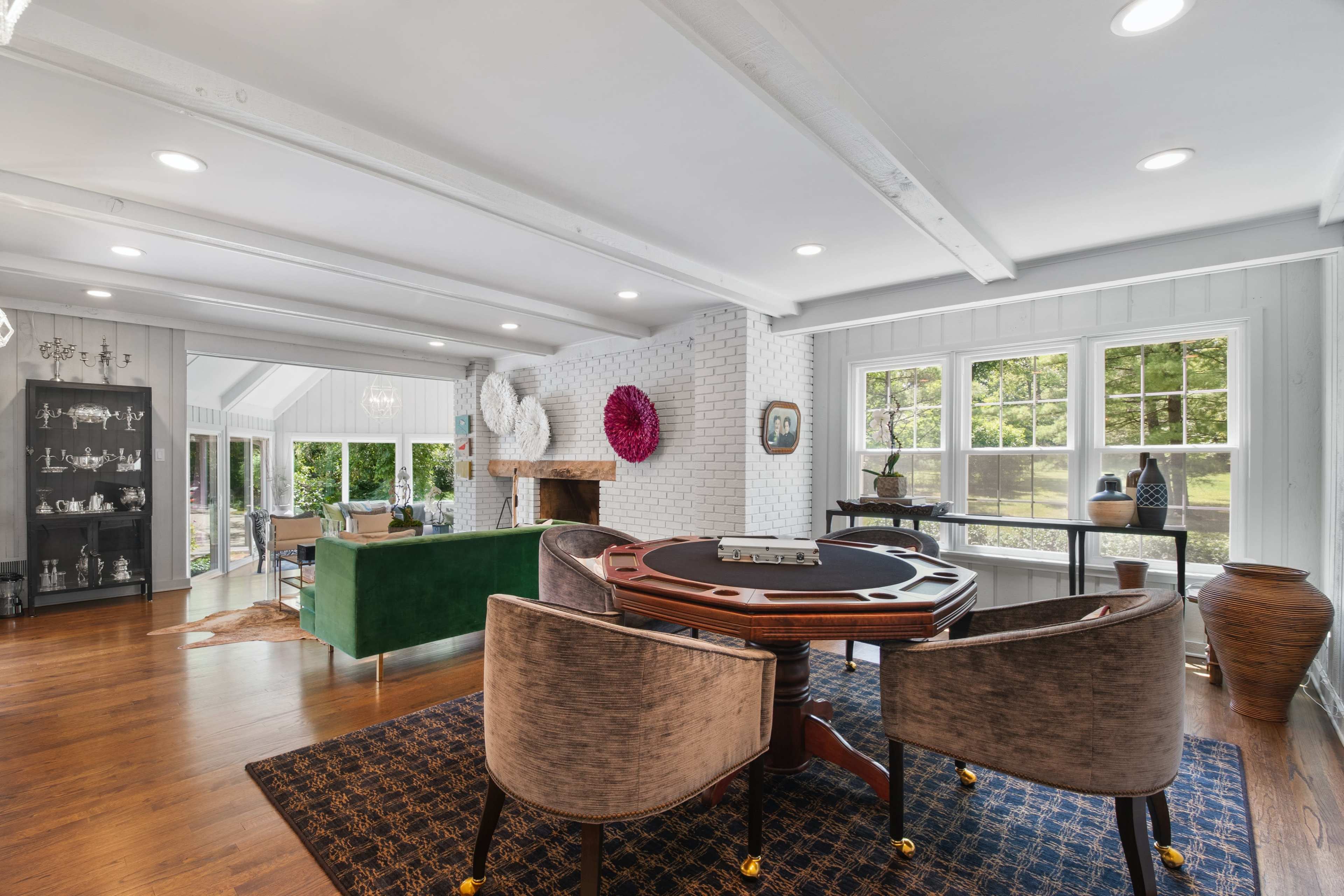 The image shows a spacious living area featuring a poker table with upholstered chairs, a green sofa, and large windows overlooking a garden.
