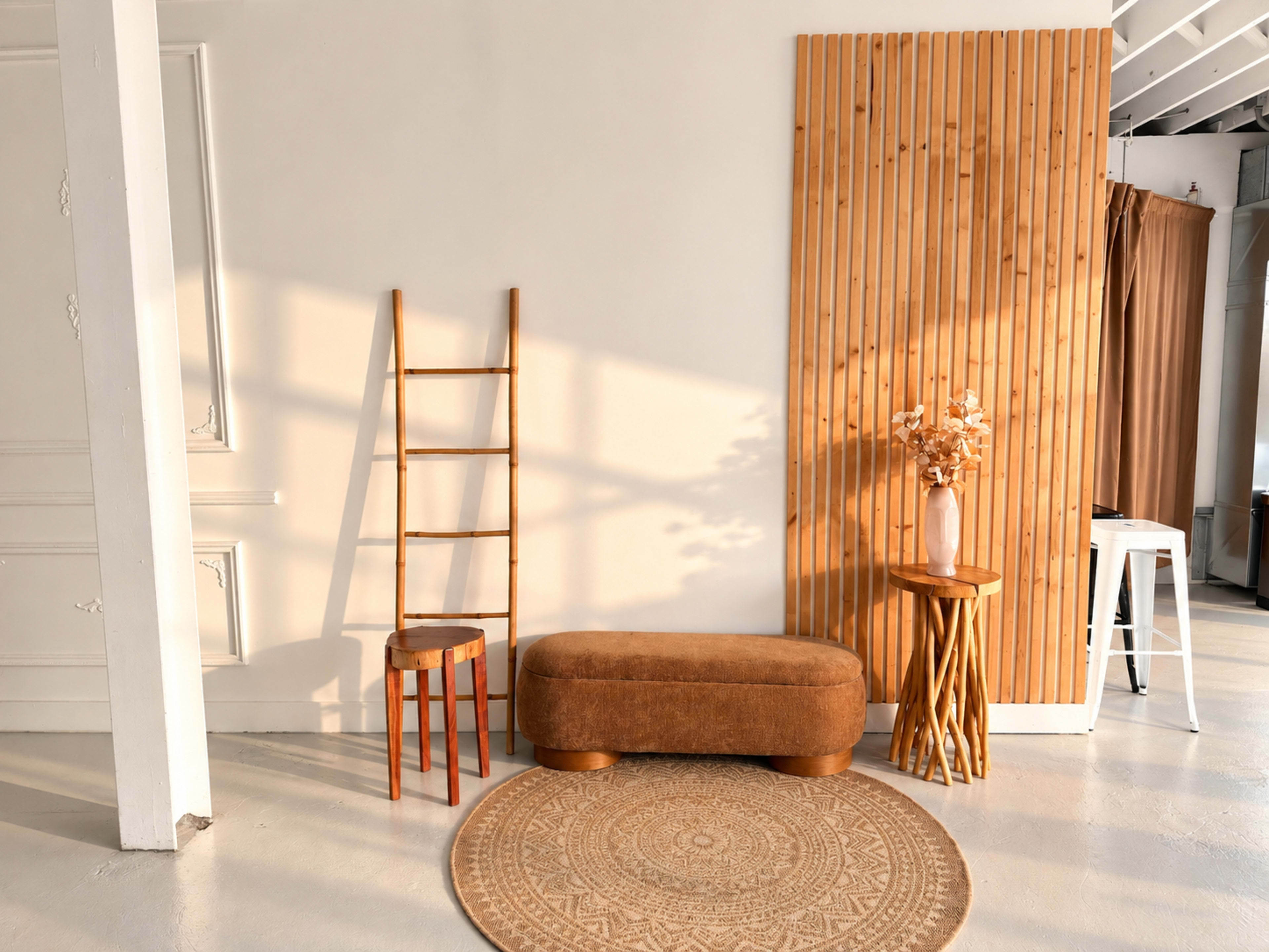 The scene features a minimalist interior with a brown ottoman, a wooden ladder, a small round table with a vase of flowers, and a textured rug on a light-colored floor.