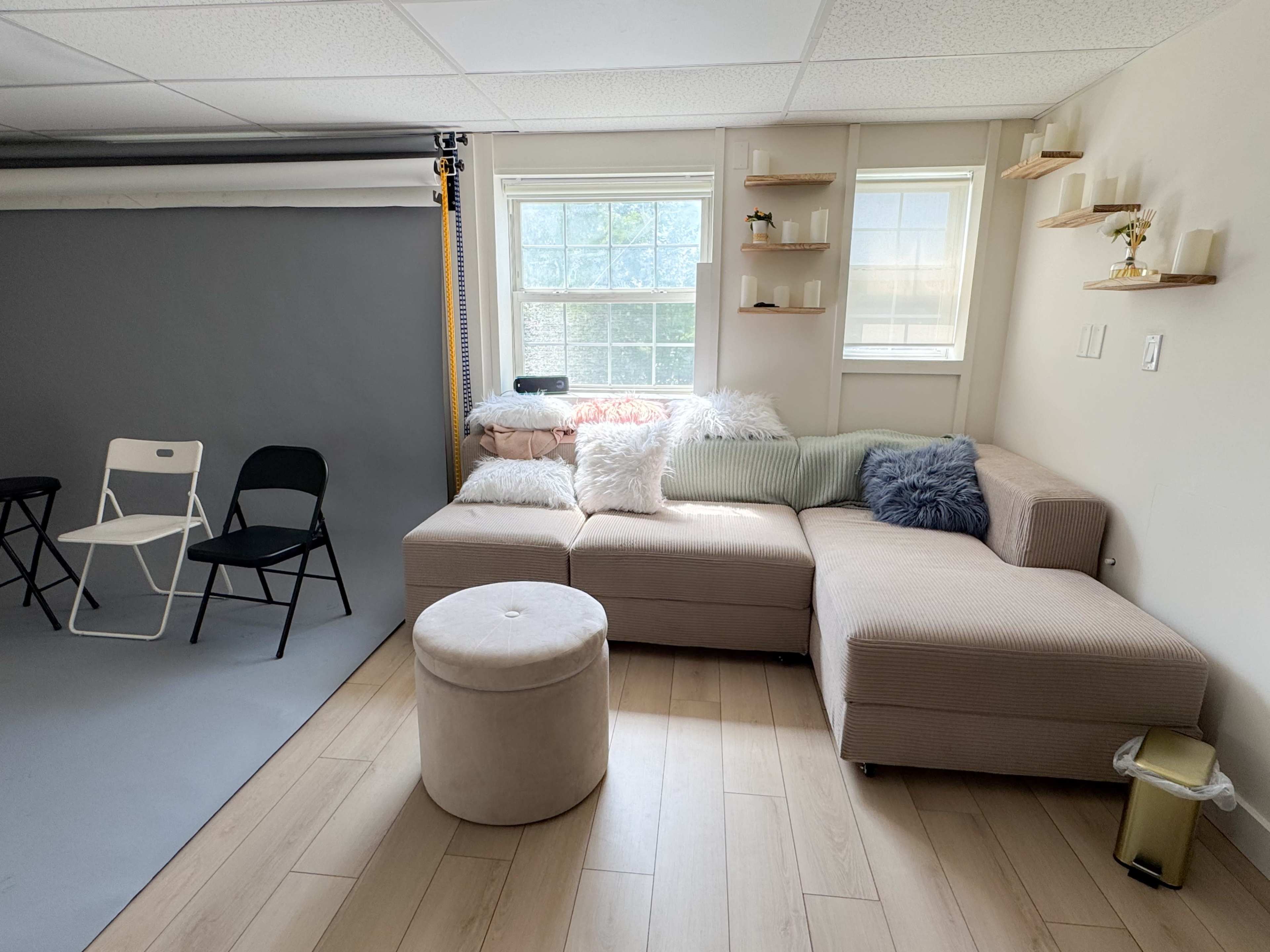 A beige sectional sofa with decorative pillows is positioned near a window in a room featuring light wood flooring and a gray backdrop.