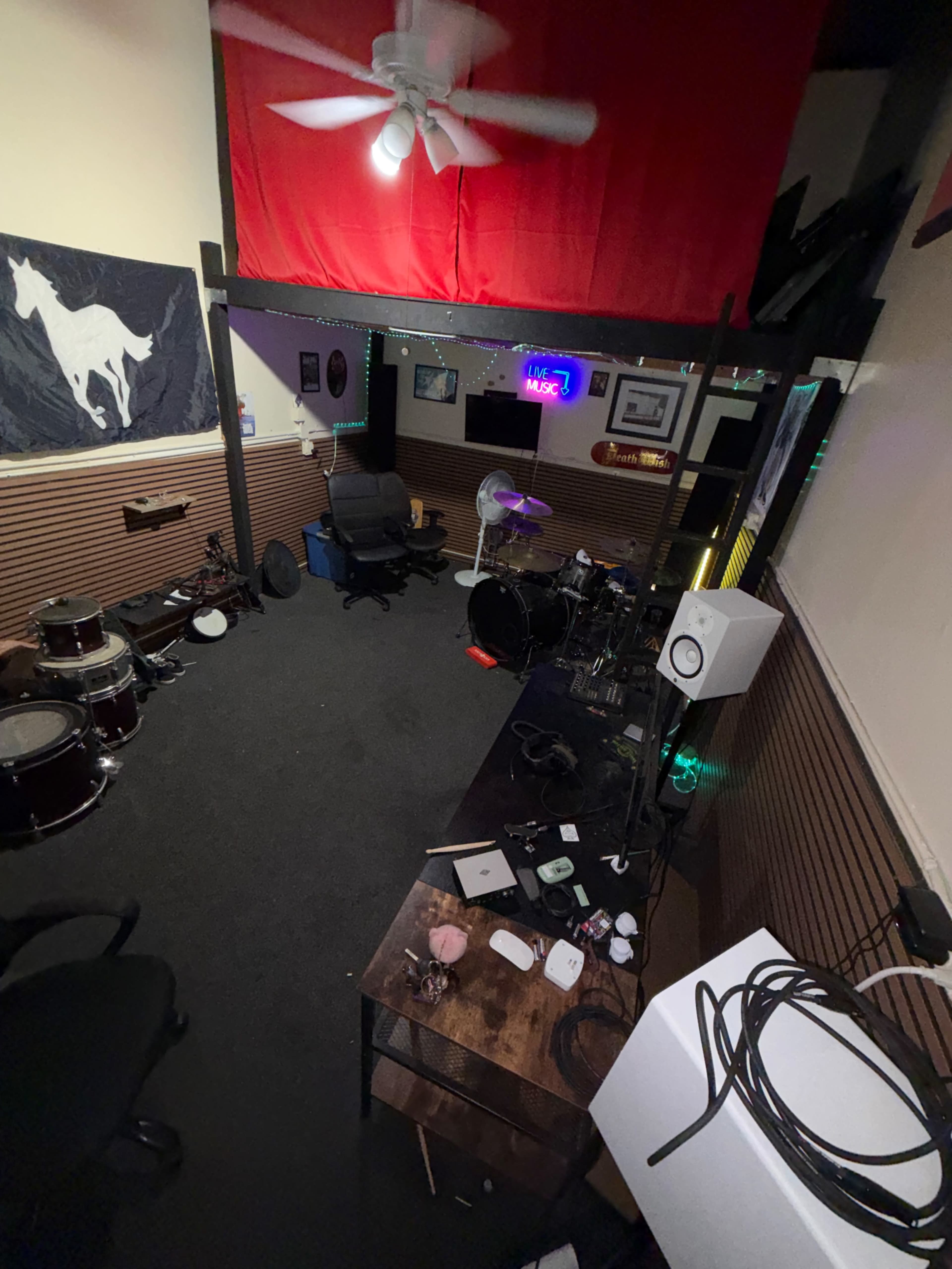 The image depicts a music rehearsal space with a drum set, a desk cluttered with equipment, and a red fabric hanging above, while a ceiling fan spins overhead.
