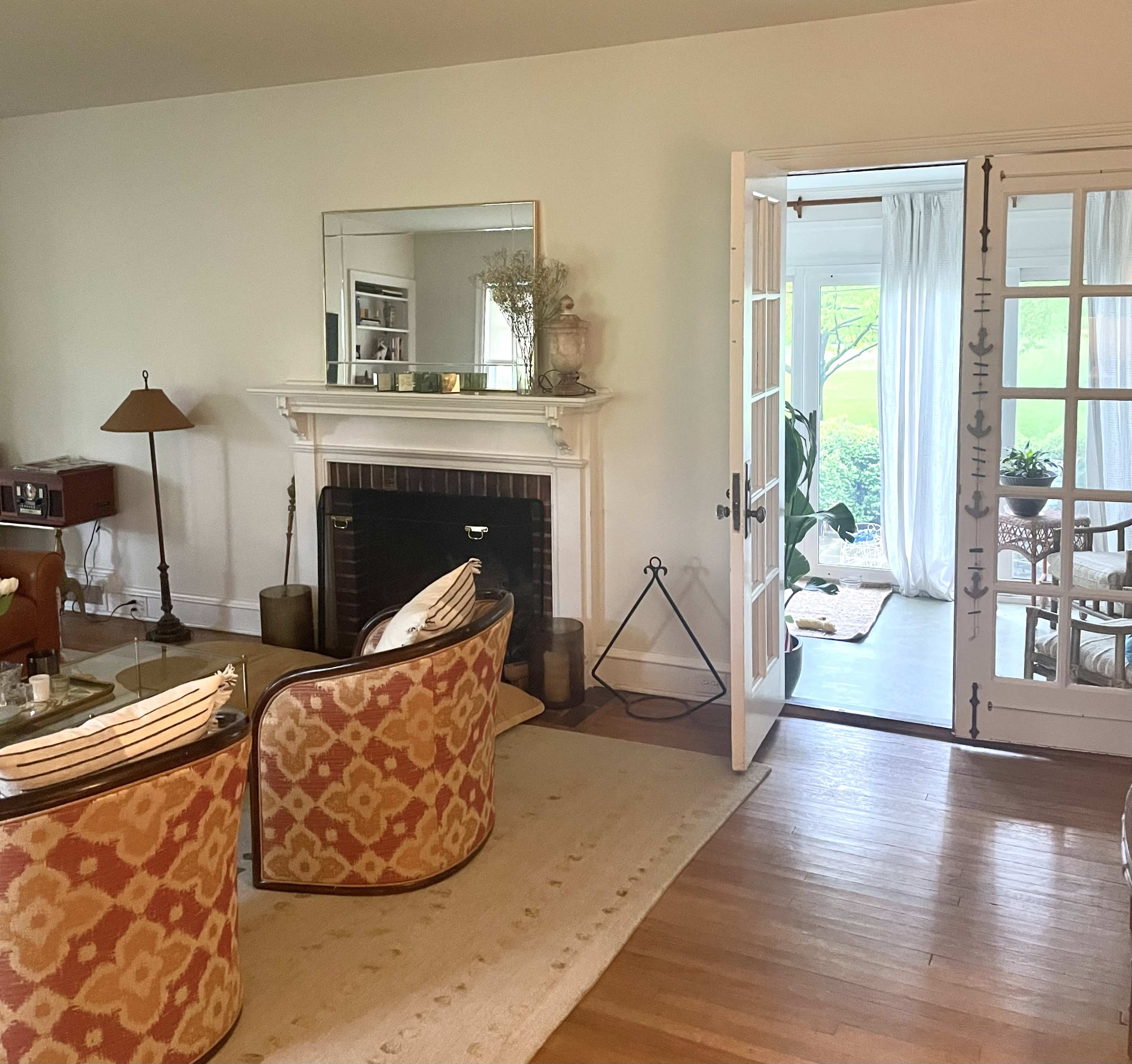 A cozy living room with patterned chairs, a fireplace, and an open doorway leading to a sunlit space with light curtains and a table.
