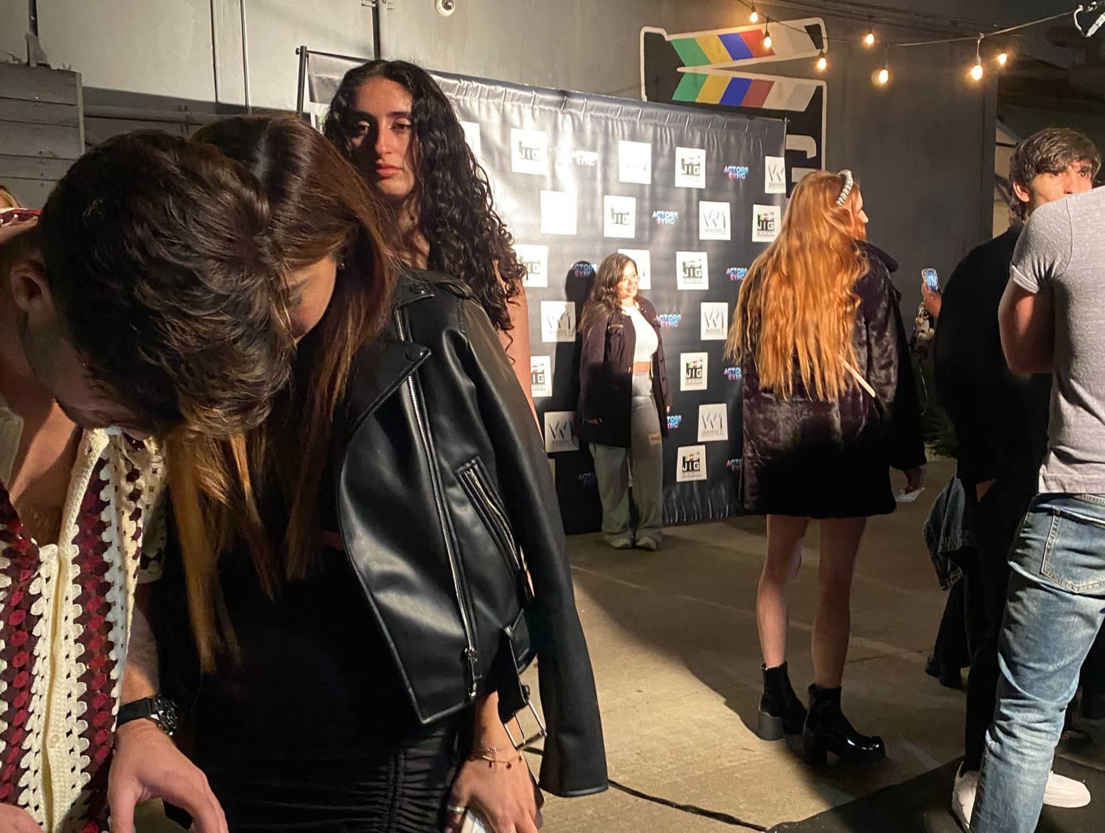 The image shows a group of people at an event, with some attendees in stylish outfits positioned near a step-and-repeat banner in the background.