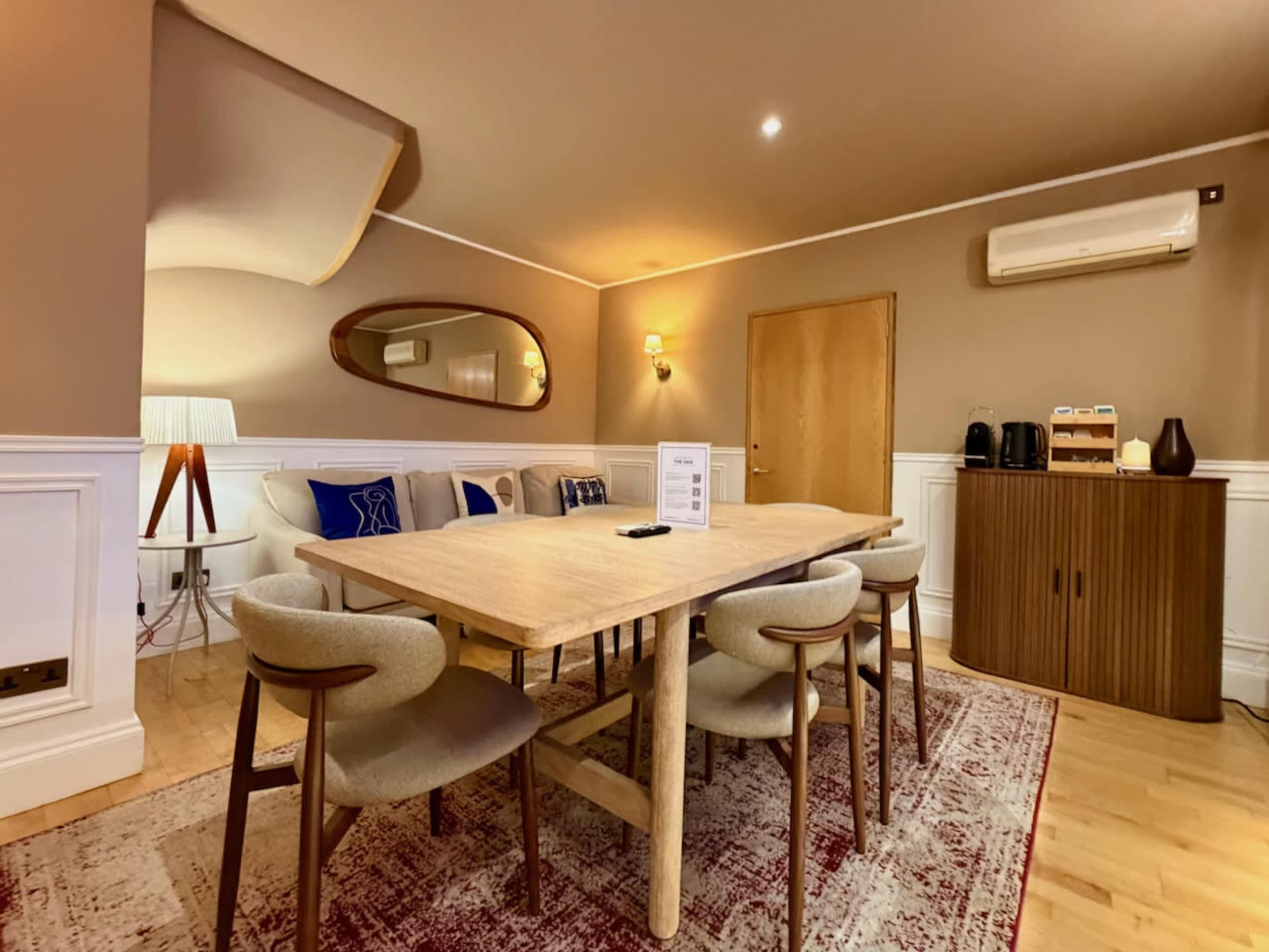 The image shows a dining area with a wooden table surrounded by four chairs, complemented by a sofa and decorative elements in a well-lit room.