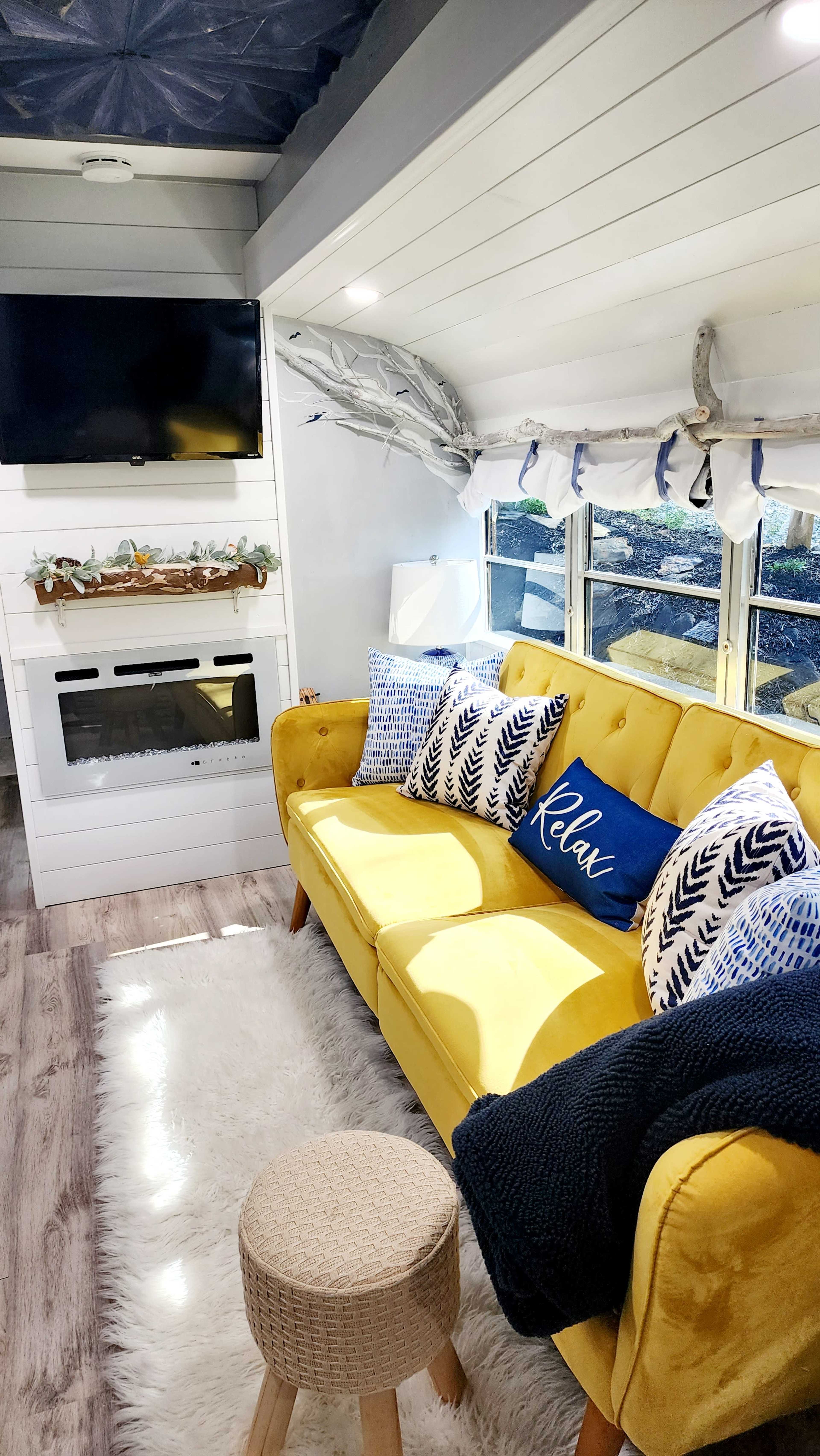 The image depicts a cozy living area with a yellow sofa adorned with decorative pillows, a television mounted on the wall, and a faux fireplace beneath a shelf, all illuminated by natural light from a window.