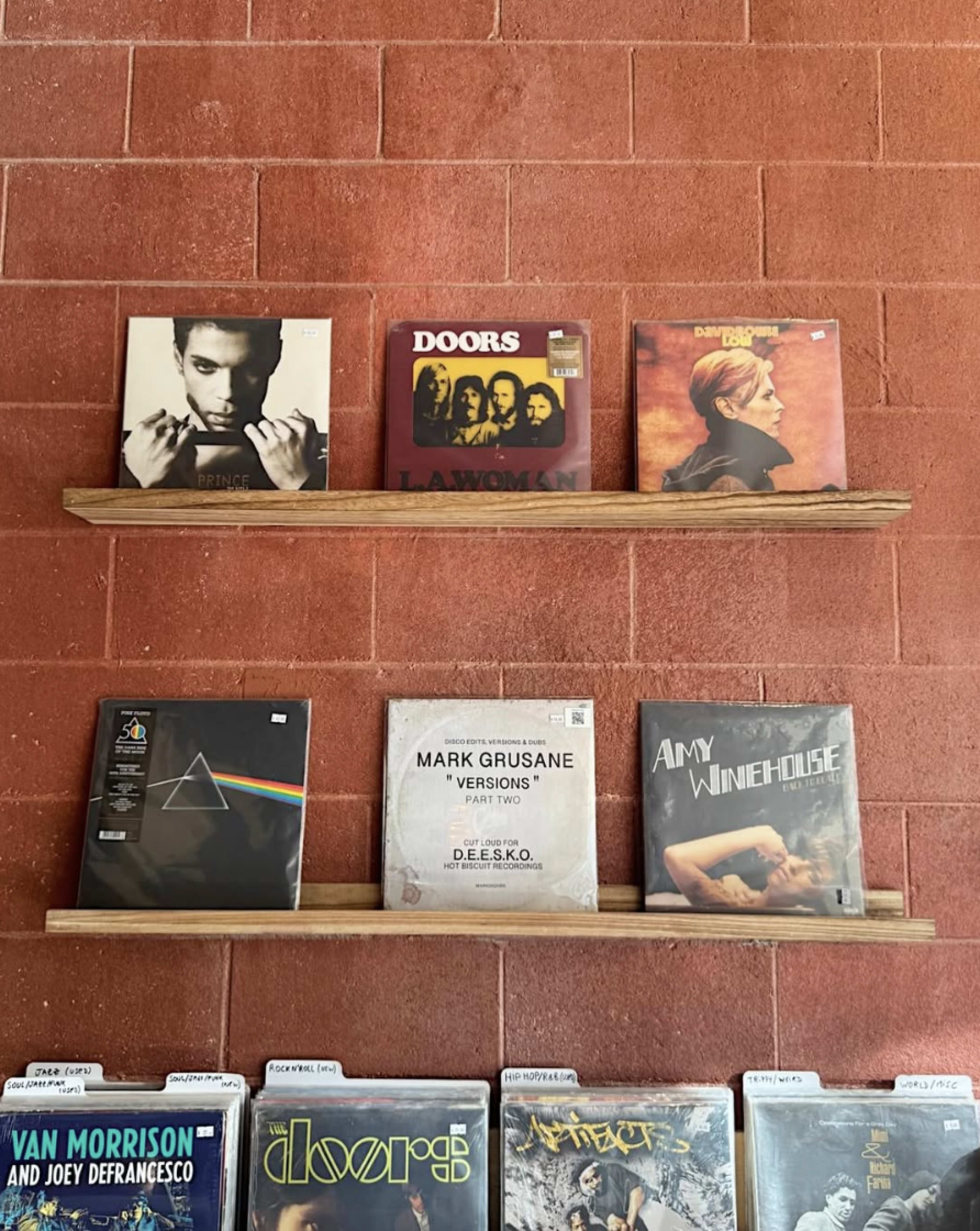 The image shows a display of vinyl records on wooden shelves against a red brick wall.