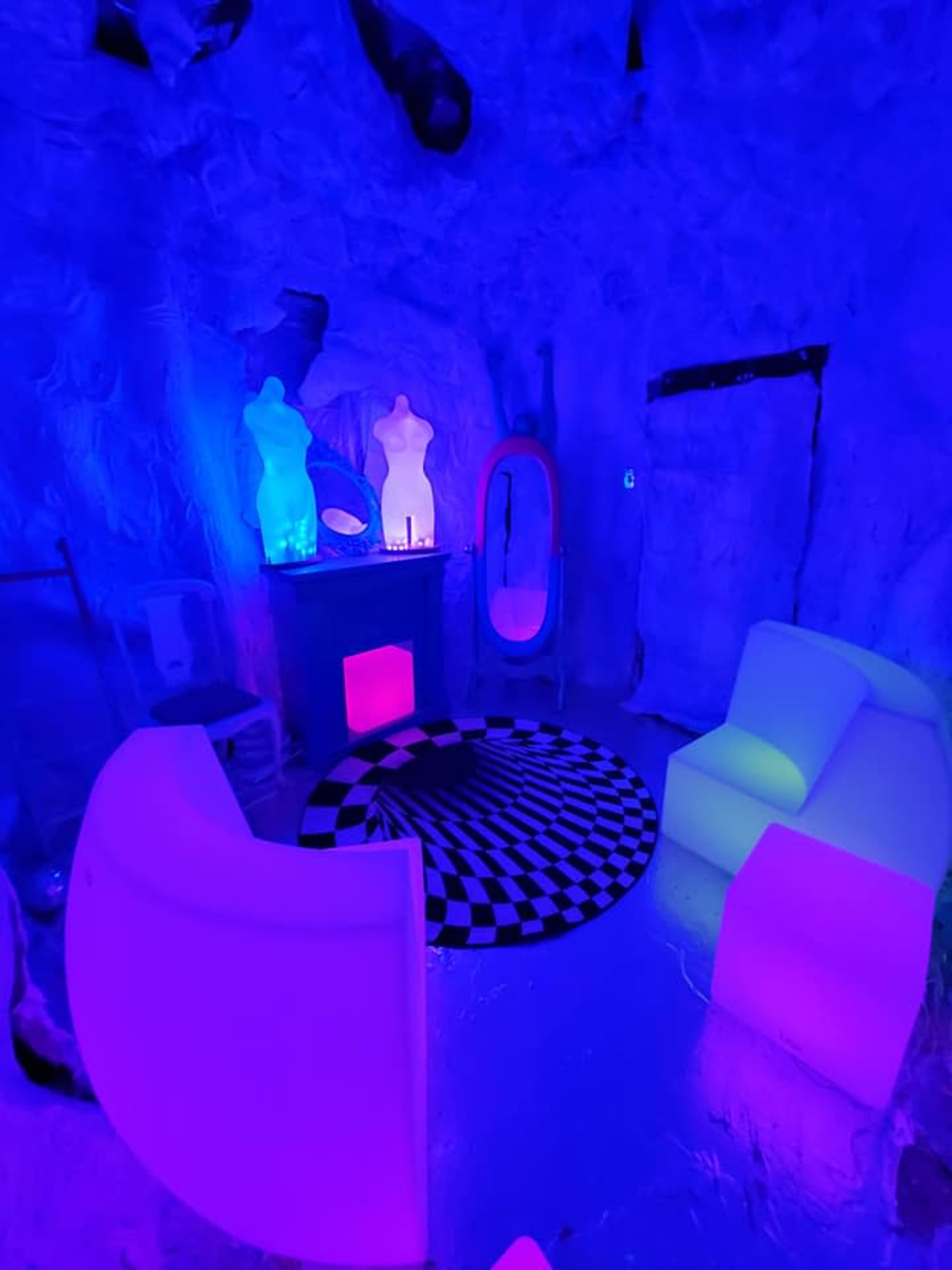 The scene features a small, dimly lit room with glowing furniture, a checkered rug, and abstract decorations illuminated by blue and purple lighting.