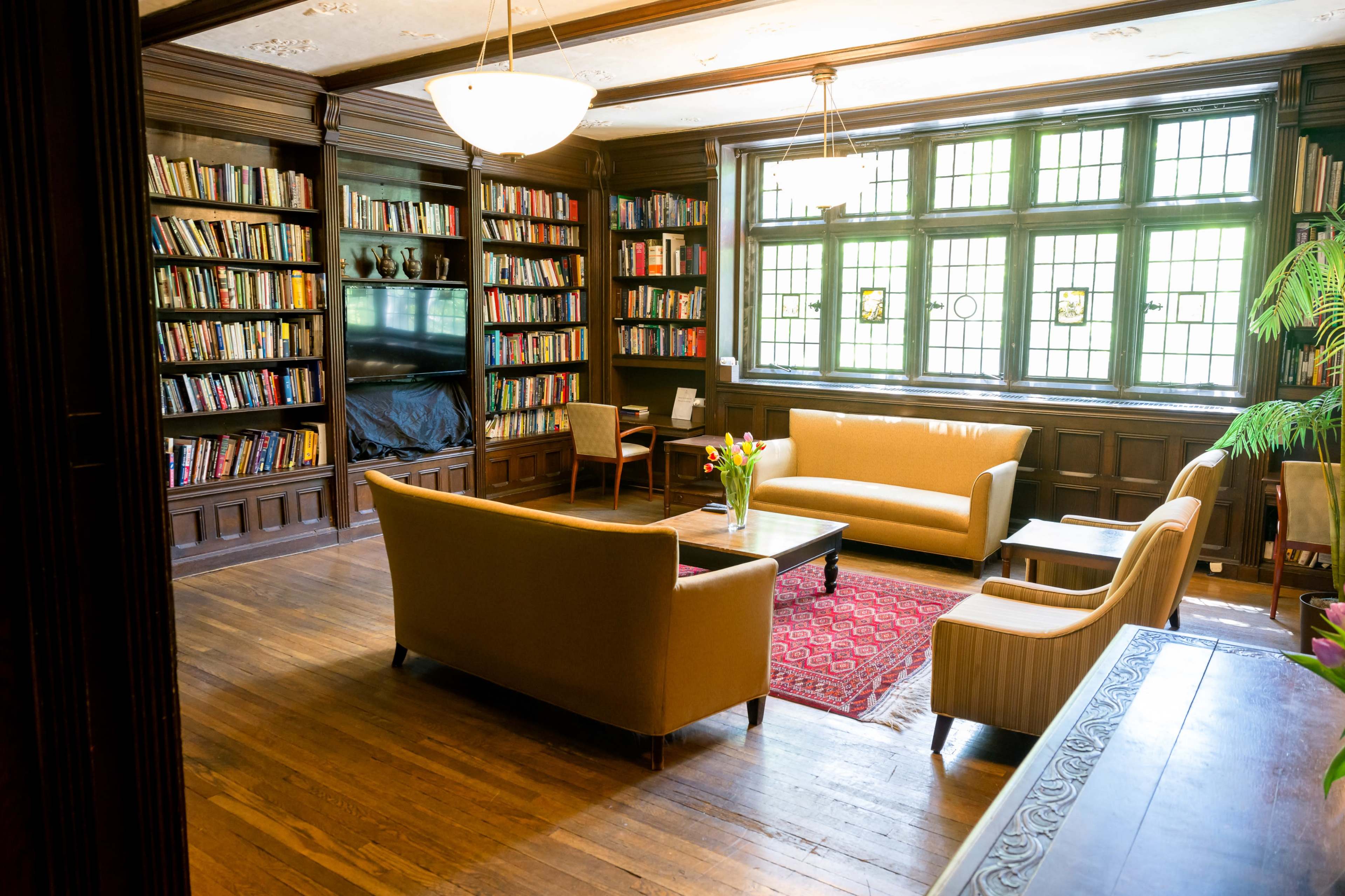 A cozy library with bookshelves lining the walls, furnished with two light-colored sofas, a coffee table, and a potted plant.
