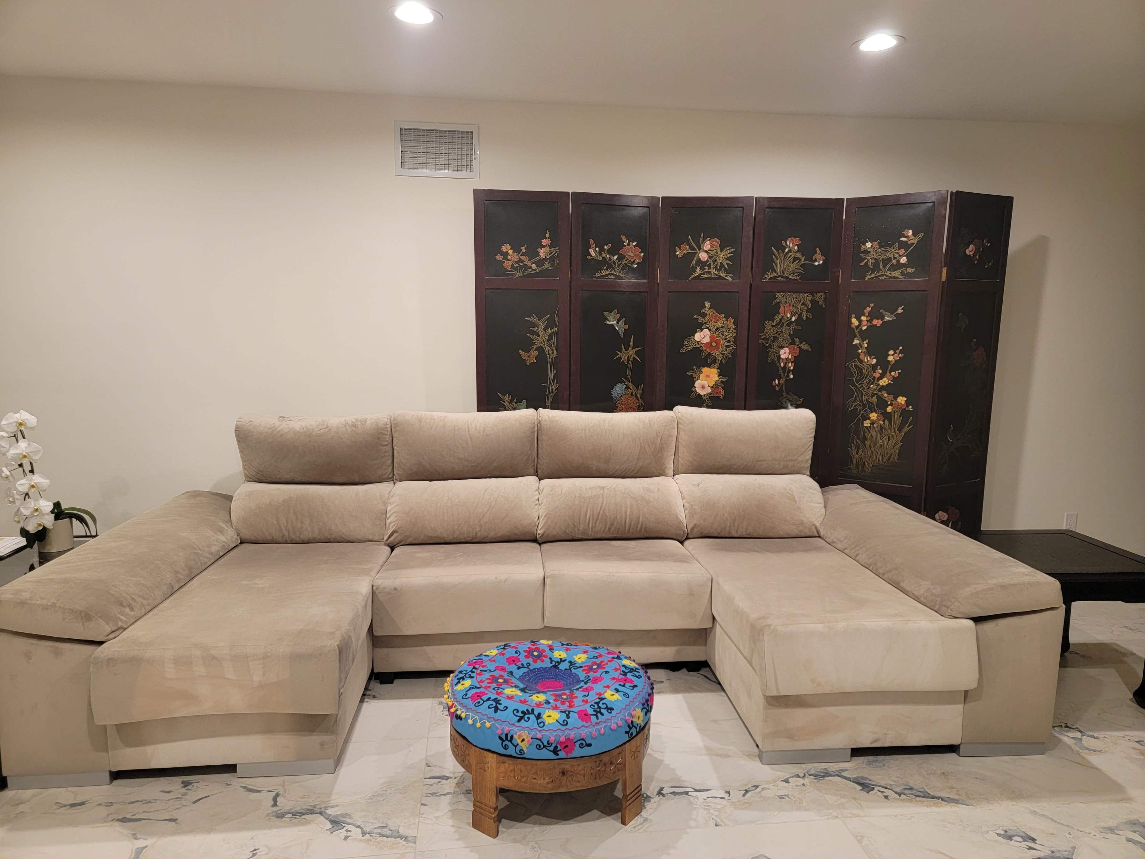 A beige sectional sofa is positioned in front of a decorative folding screen adorned with floral designs, alongside a round, colorful table with a floral pattern.