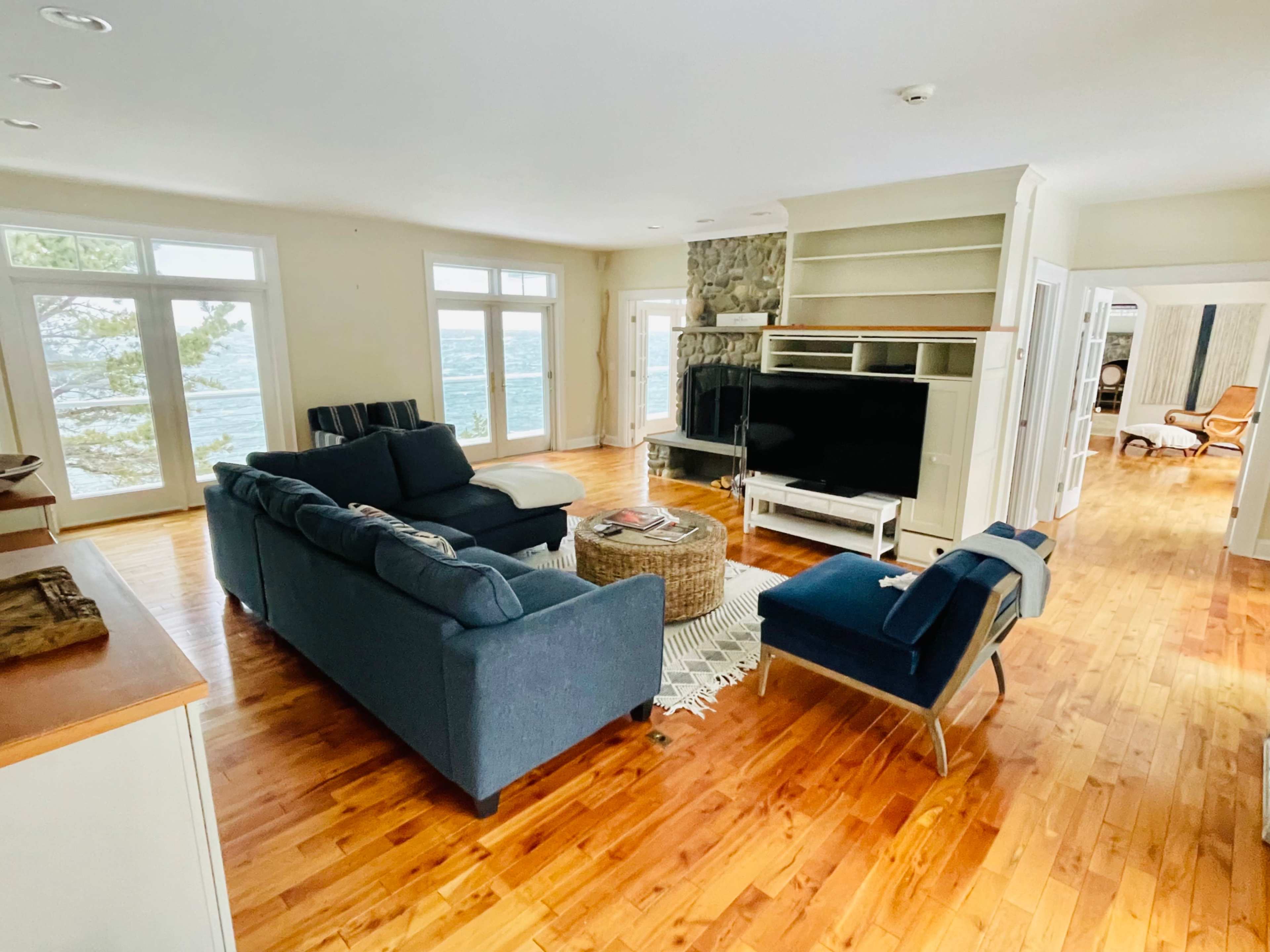 The living room features a blue sectional sofa, a coffee table, and a stone fireplace, with large windows overlooking a body of water.