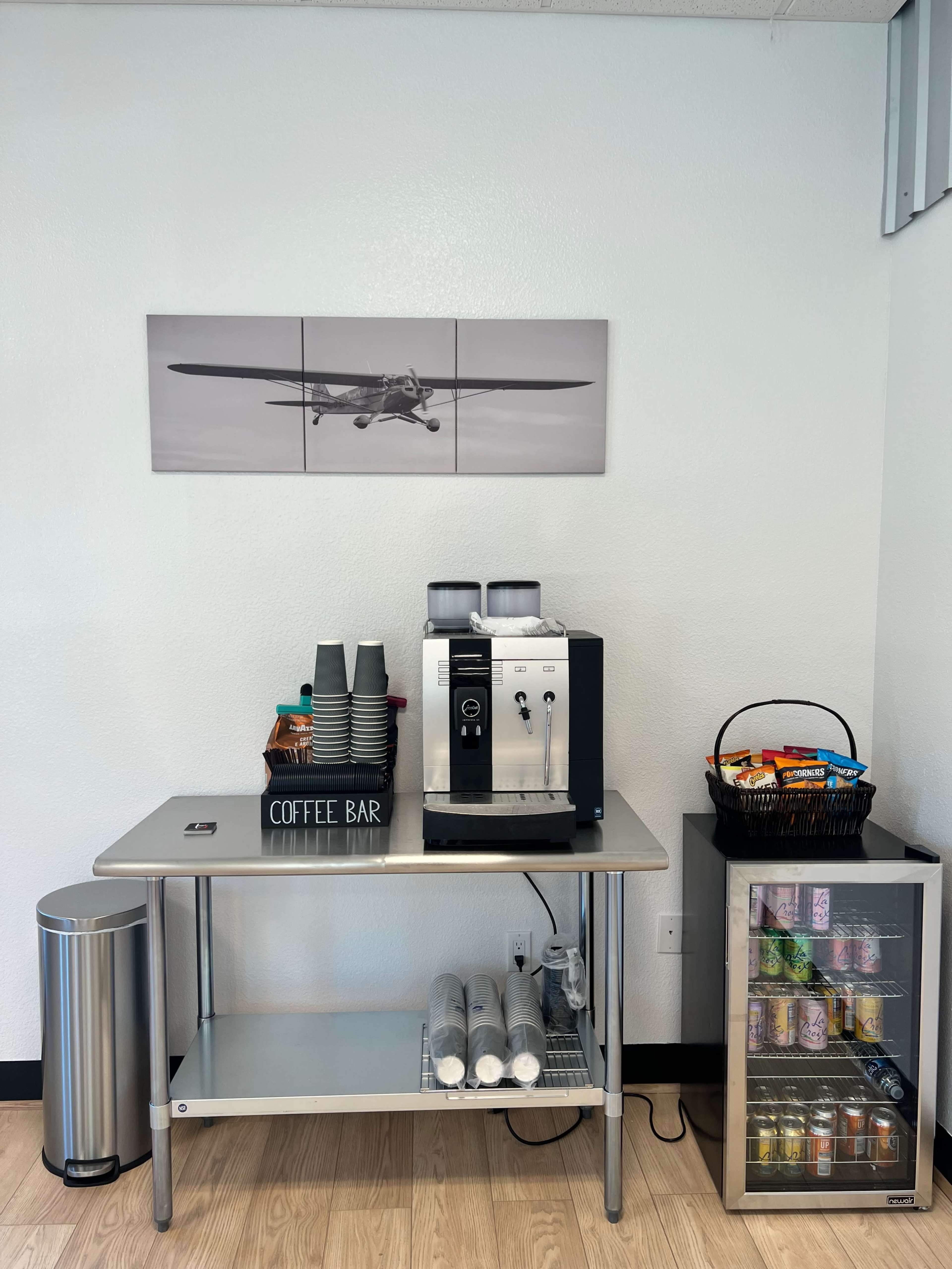 A coffee bar setup with a coffee machine, stacked cups, a waste bin, and a glass-front refrigerator displaying various snacks and drinks.