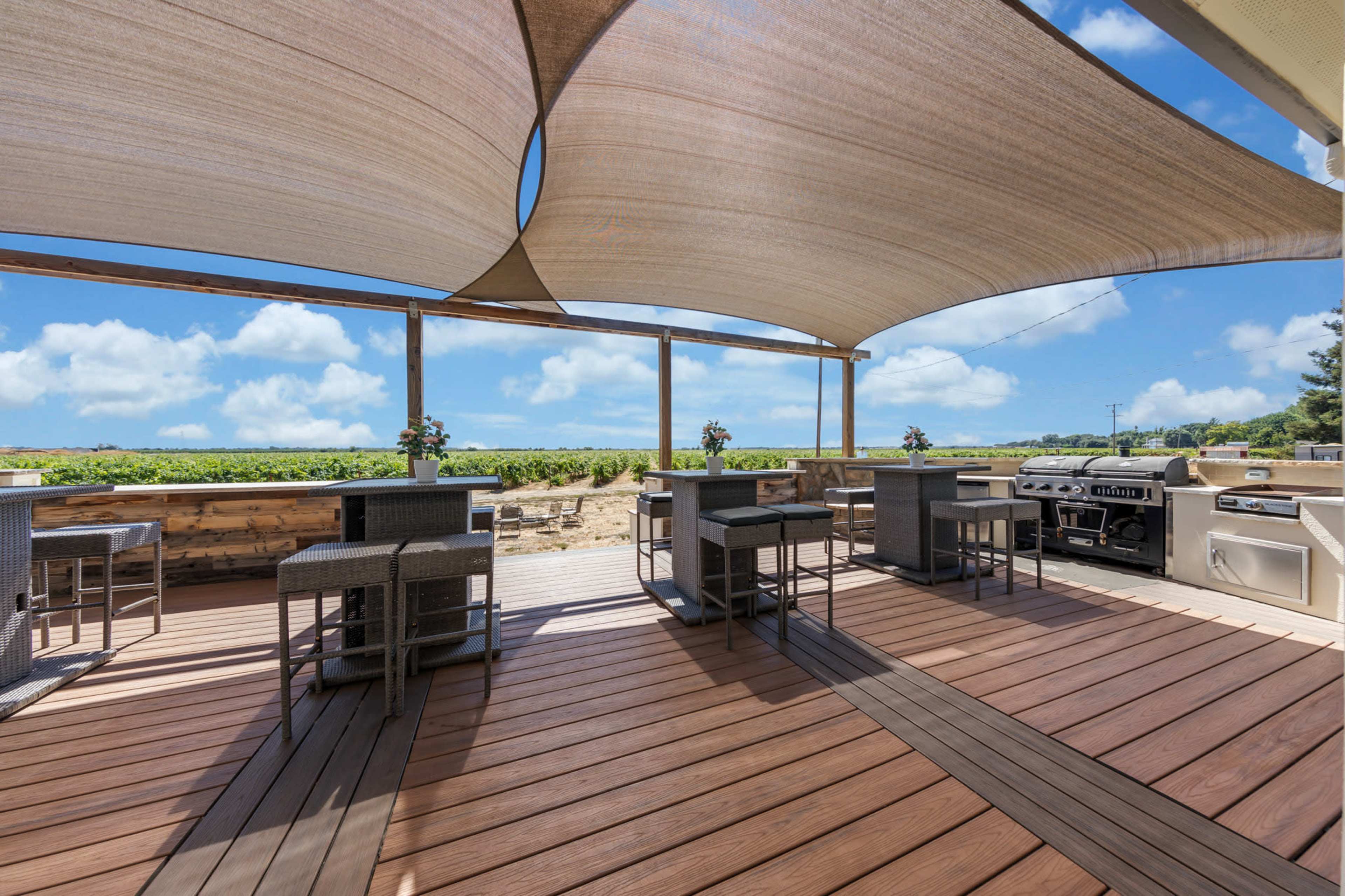 A spacious outdoor patio with shaded seating areas, a grill, and a view of a vineyard under a clear blue sky.