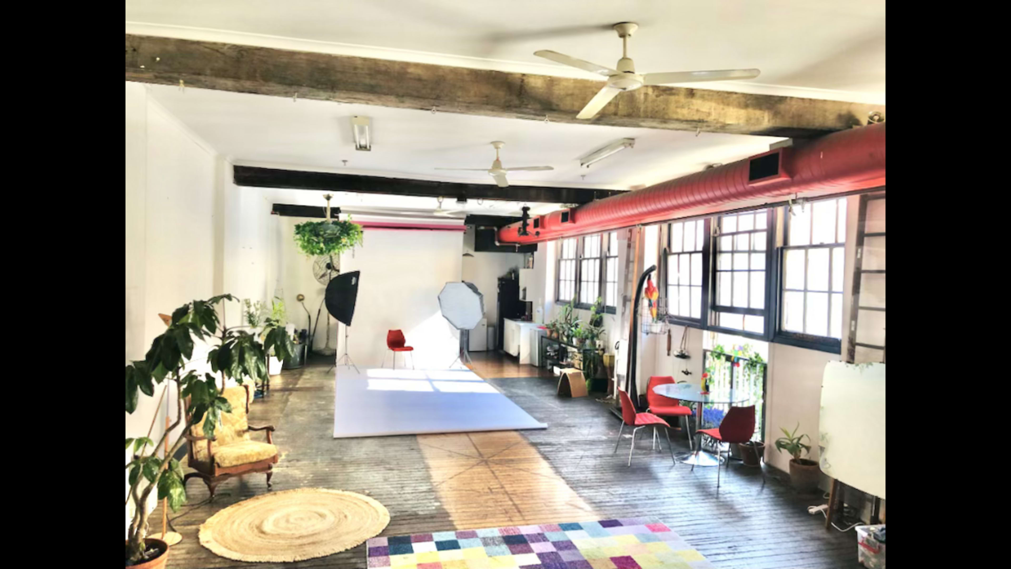 The image shows a spacious, well-lit studio featuring wooden floors, large windows, plants, and various seating areas, including a red chair and a rug.