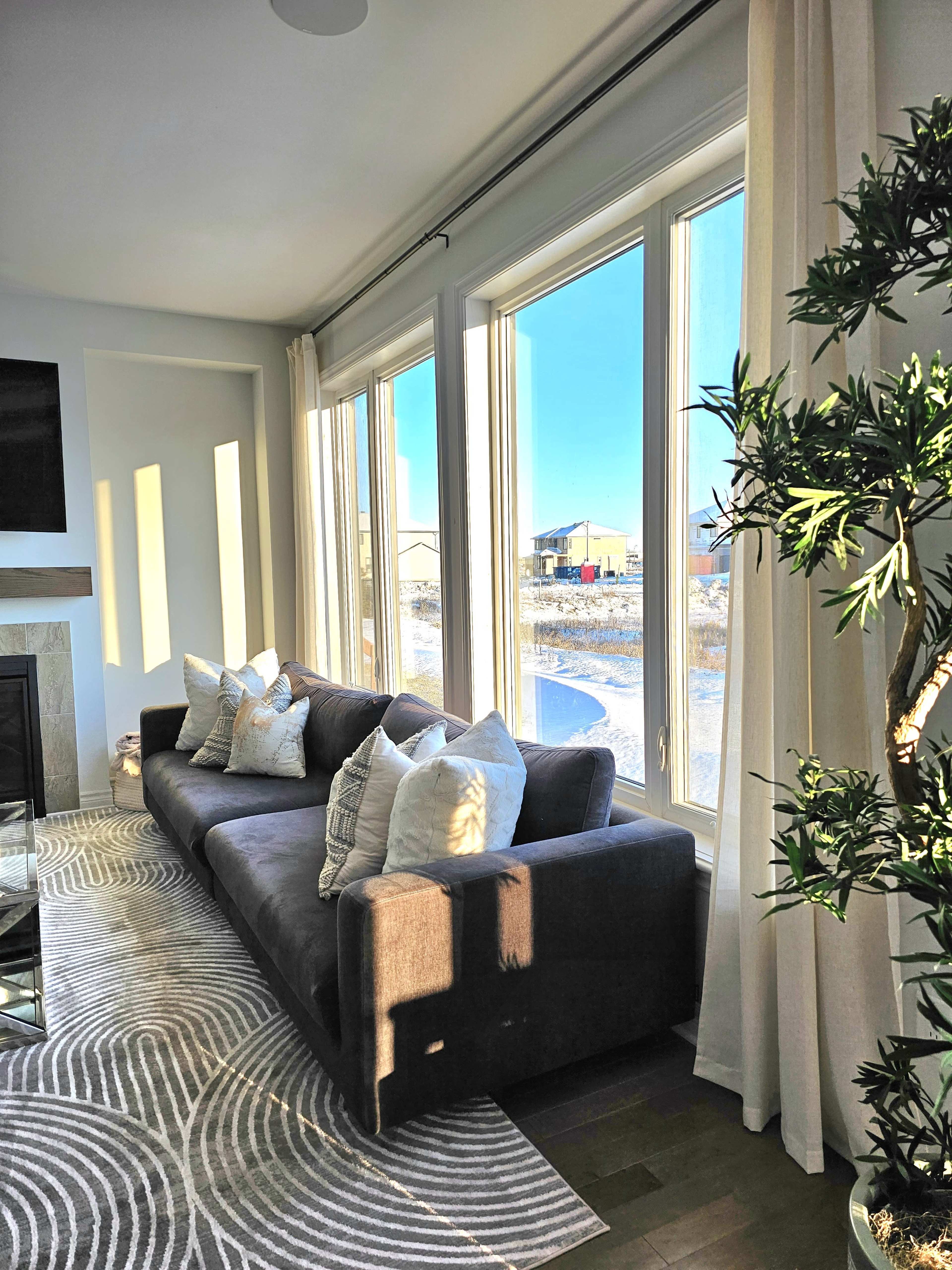 A gray sofa with decorative pillows is positioned next to large windows that let in natural light, overlooking a view of a winter landscape.