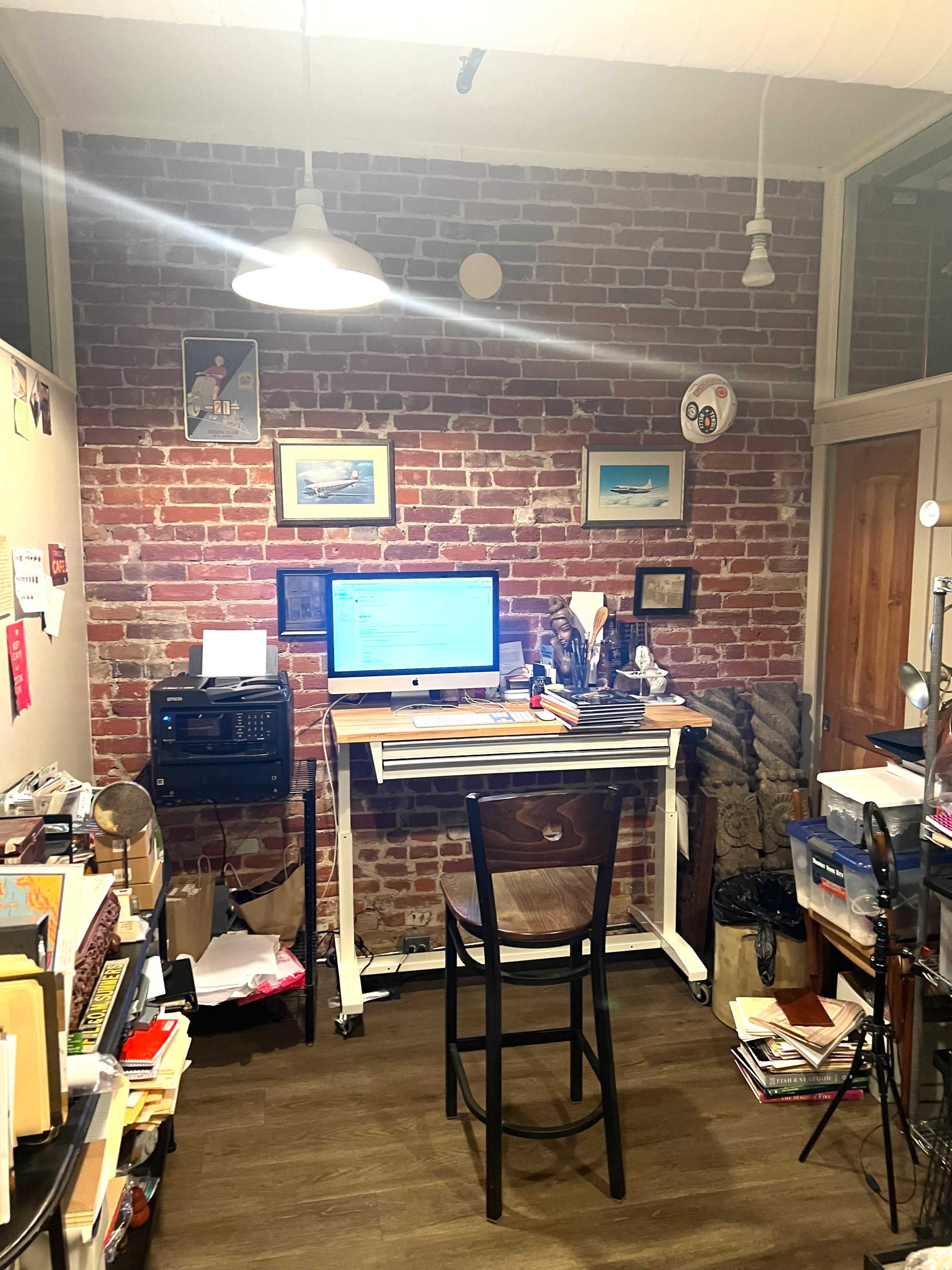 A home office with a desk, computer, and various items scattered around, against a backdrop of exposed brick walls.