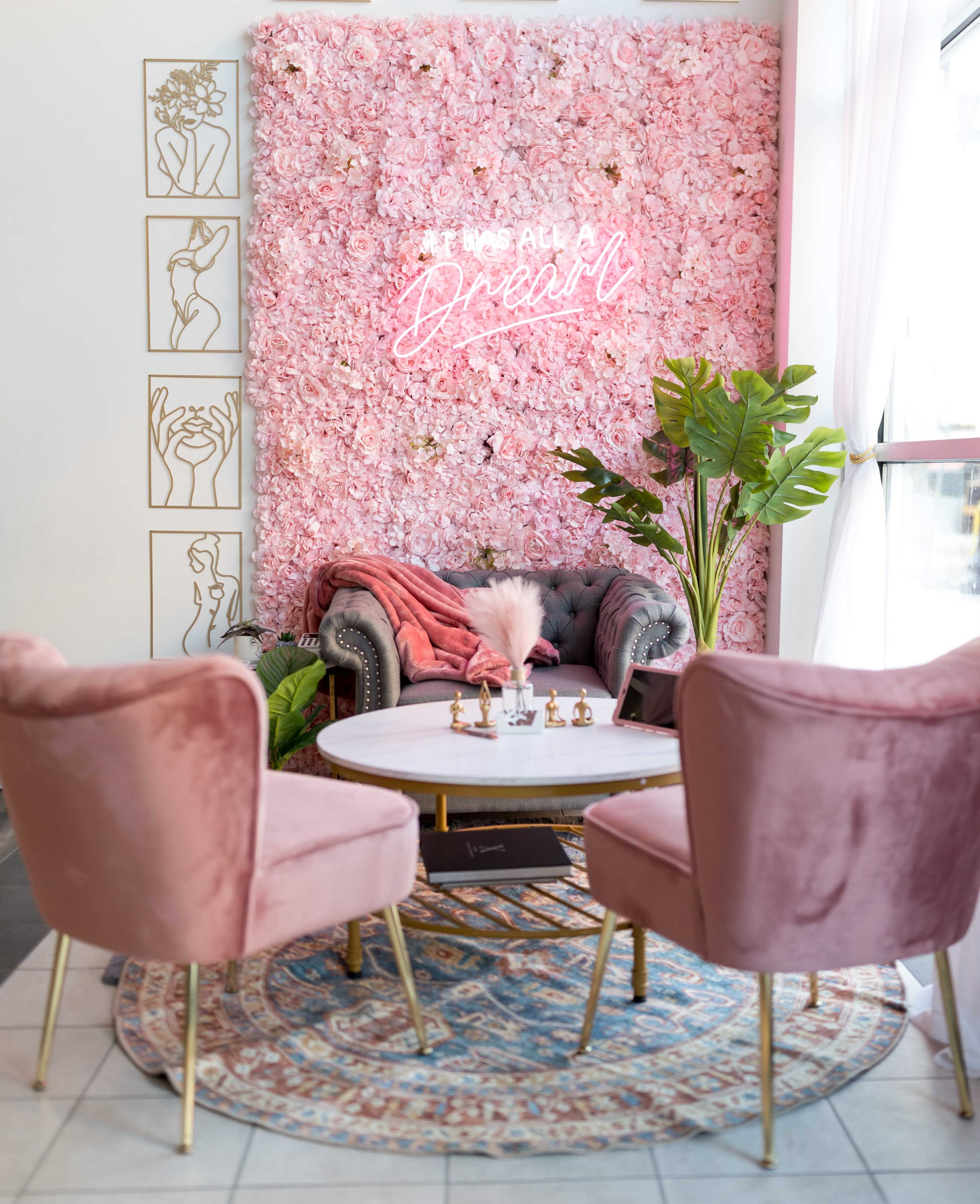 A cozy lounge area features a pink floral wall, a gray velvet sofa, two pink armchairs, and a round coffee table on a patterned rug.