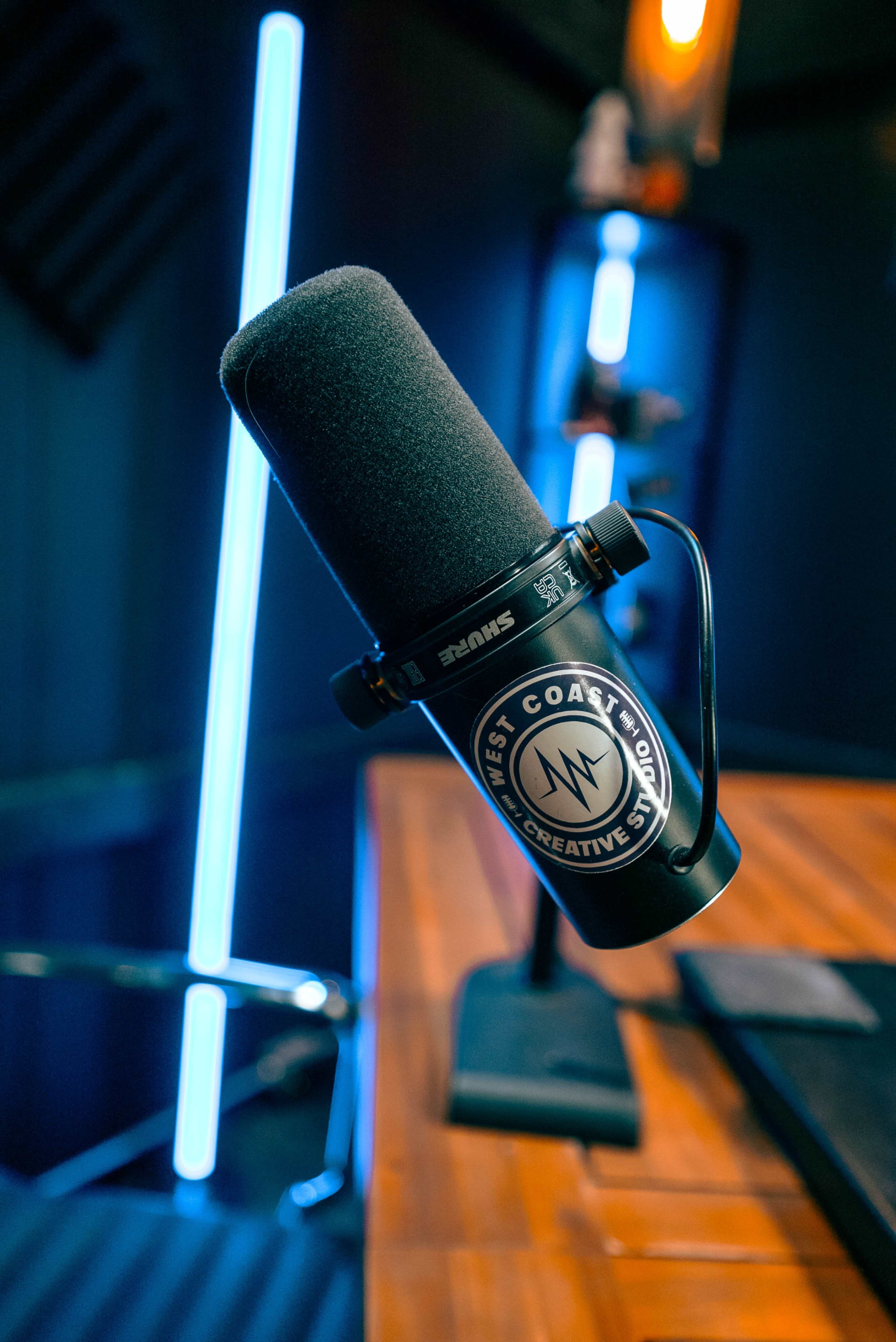 A close-up of a black microphone mounted on a wooden table, featuring a logo for "West Coast Creative" and surrounded by blue LED lighting in a studio setting.