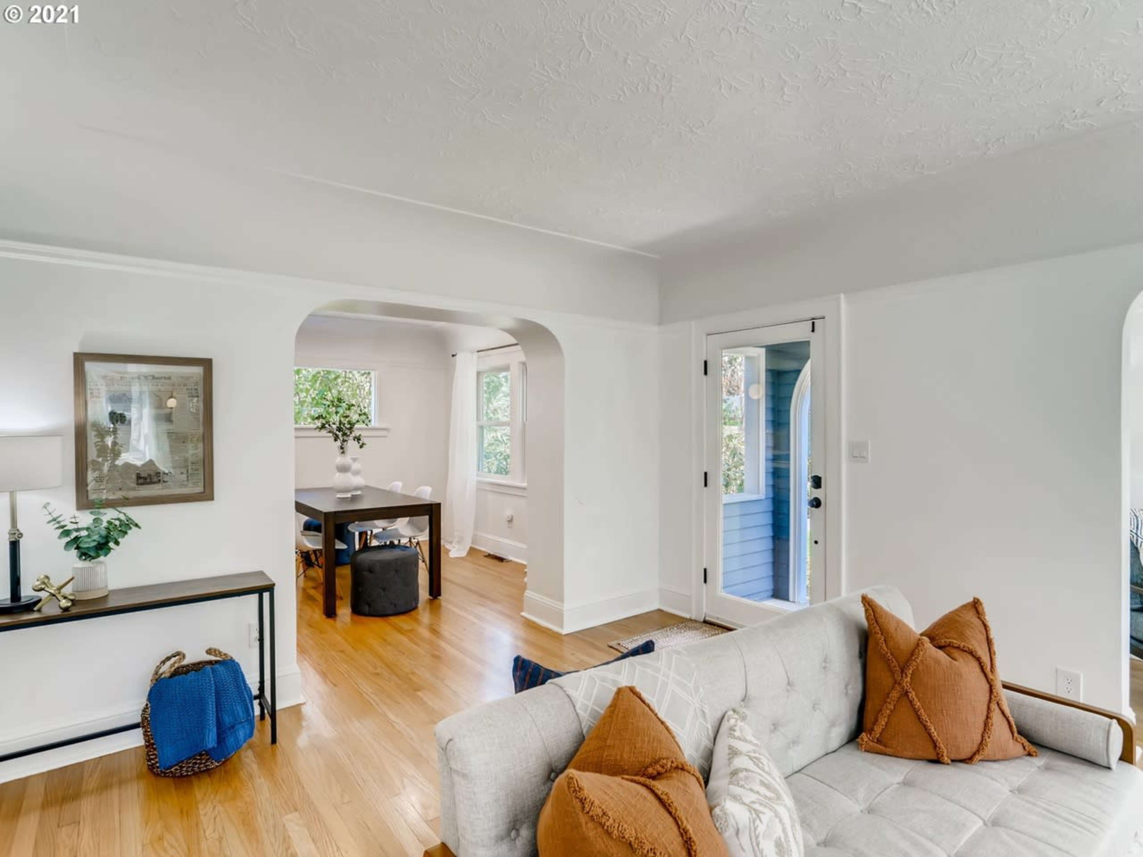 The image shows a bright living room with a light-colored sofa, decorative pillows, and a dining area visible through an archway.