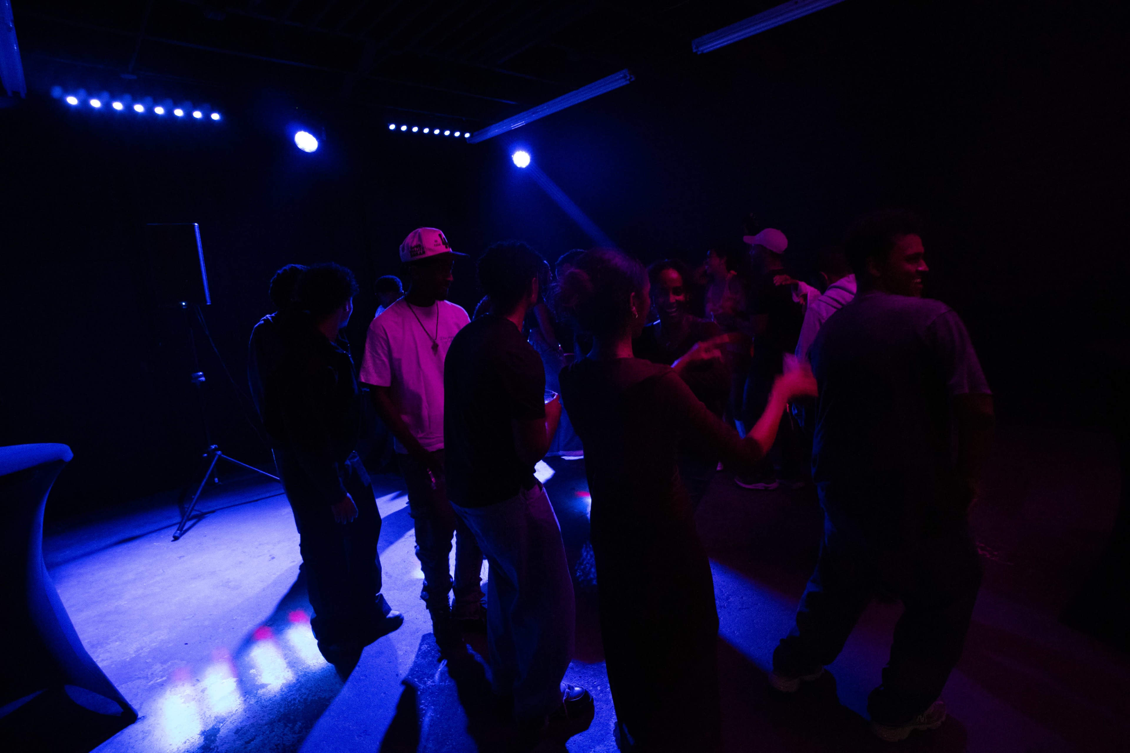 A group of people is gathered in a dimly lit room with blue lighting, engaging in conversation and movement.