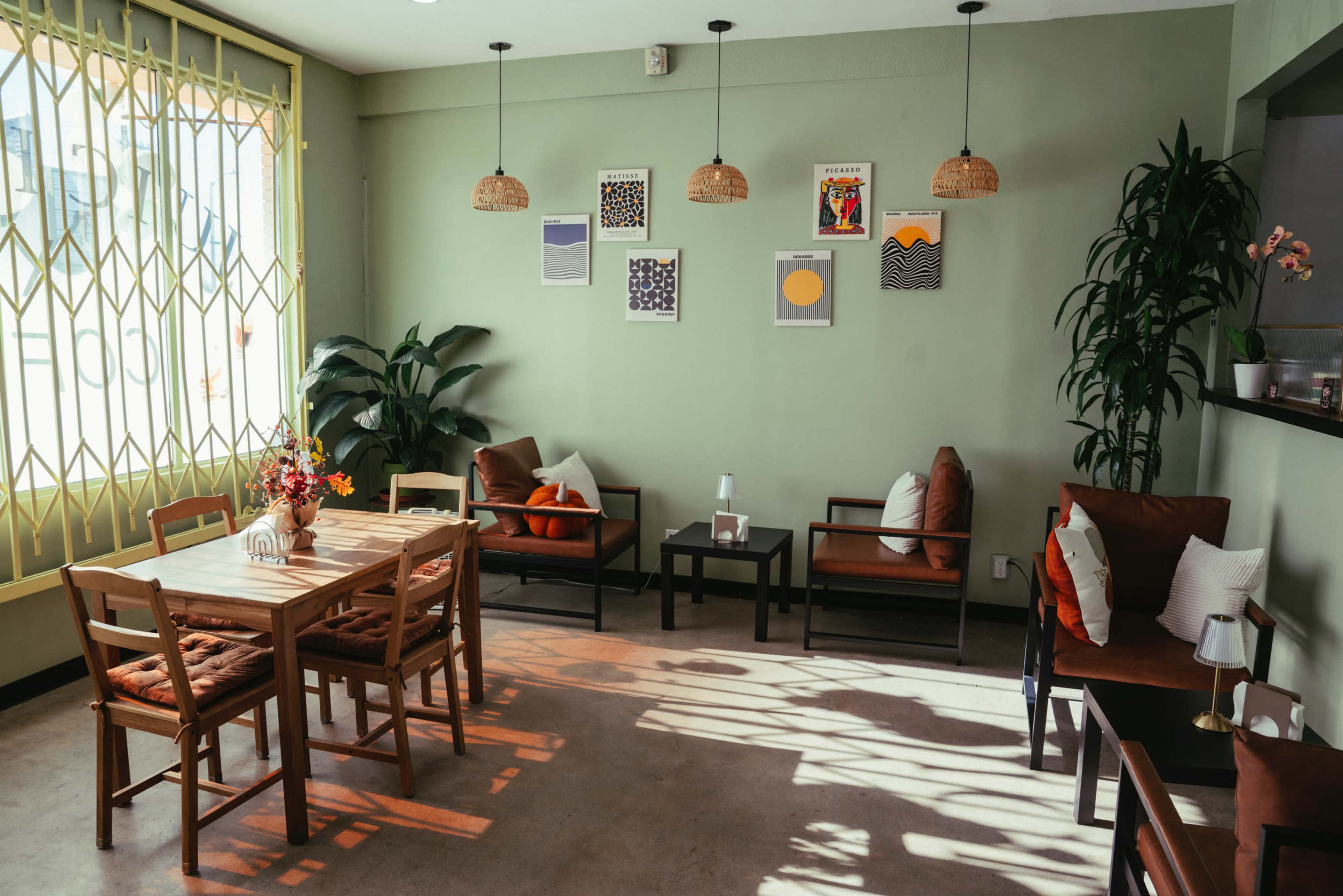 The image shows a cozy café interior with a wooden table and chairs, surrounded by plants and adorned with wall art, under soft illumination from hanging lights.