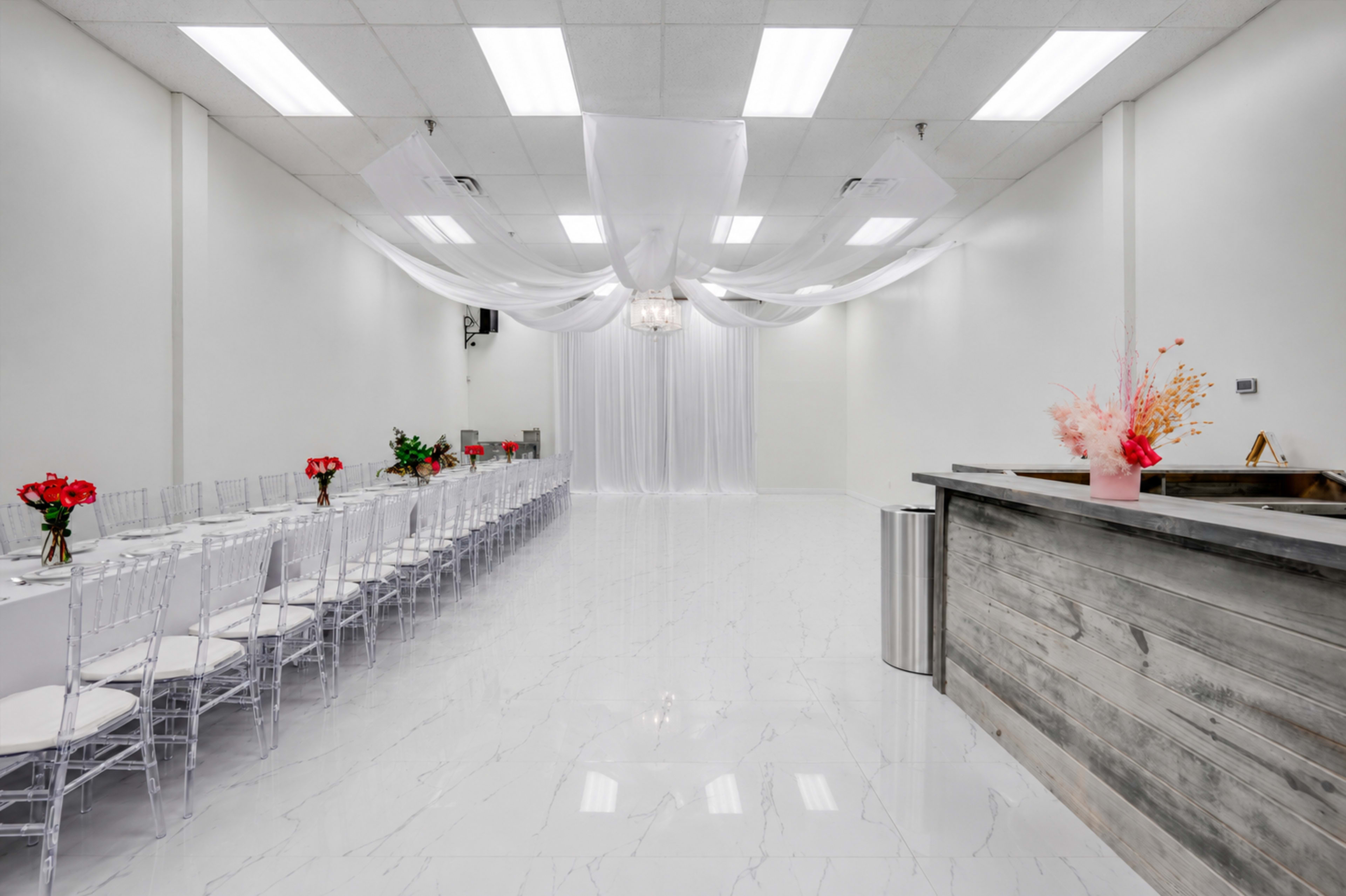 The image shows a spacious event room with a long table set for dining, decorated with flowers, and a bar area in one corner under suspended drapery.