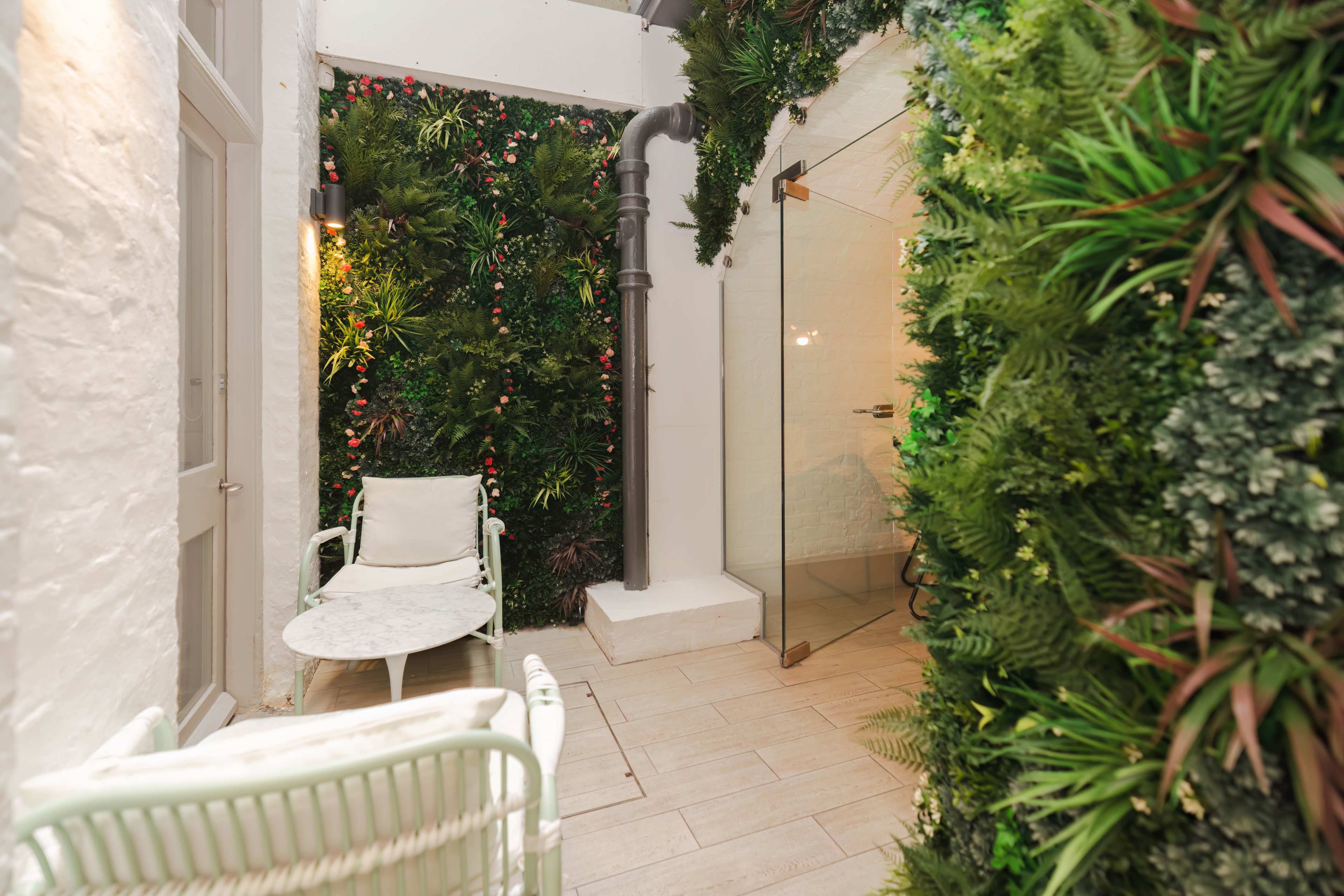 A small indoor space with greenery-covered walls, a glass shower enclosure, and a seated area featuring a white chair and a round table.