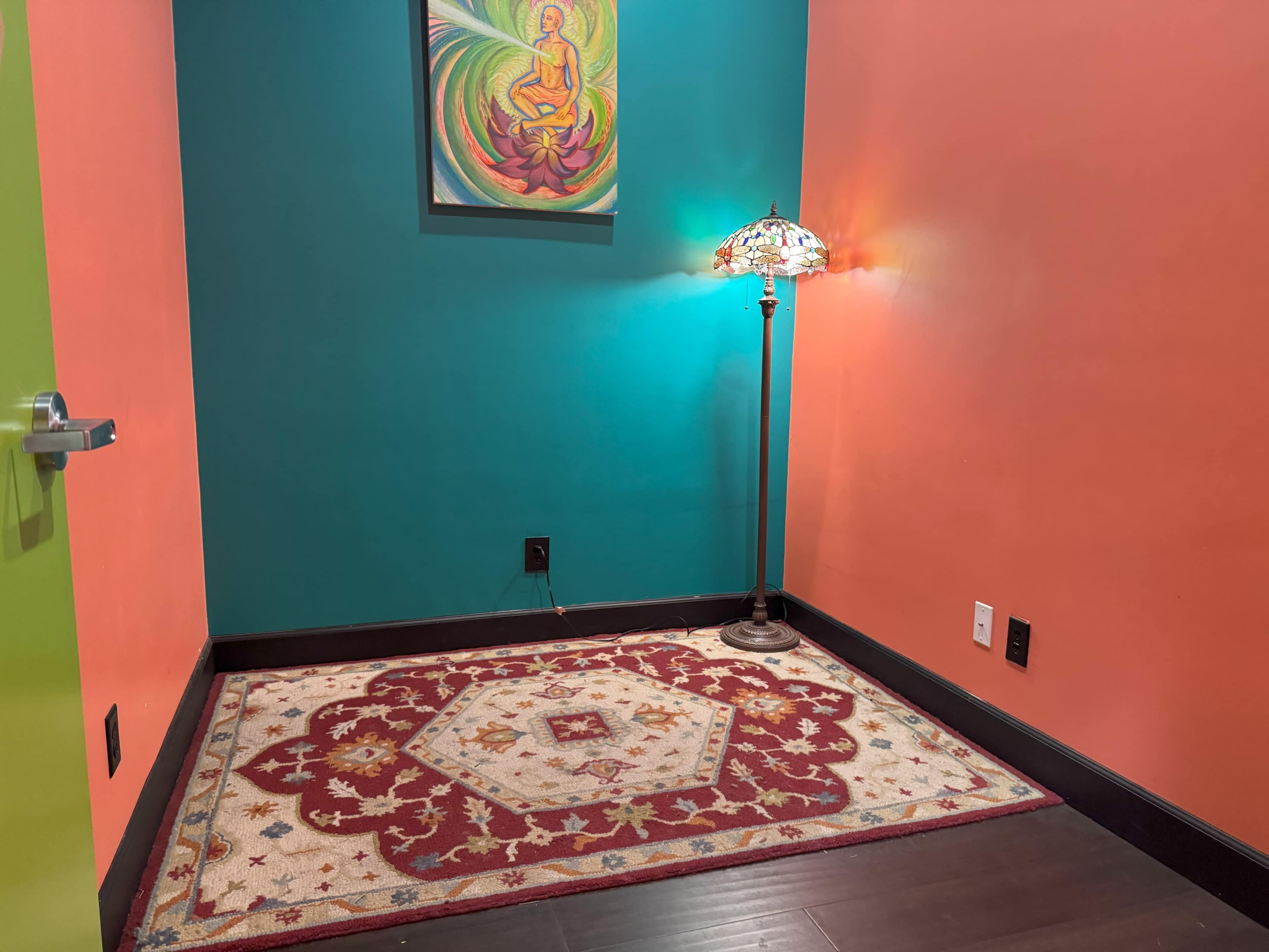 The image shows a small corner of a room with an intricate rug, a decorative lamp, and a colorful mural with a circular design on the wall.