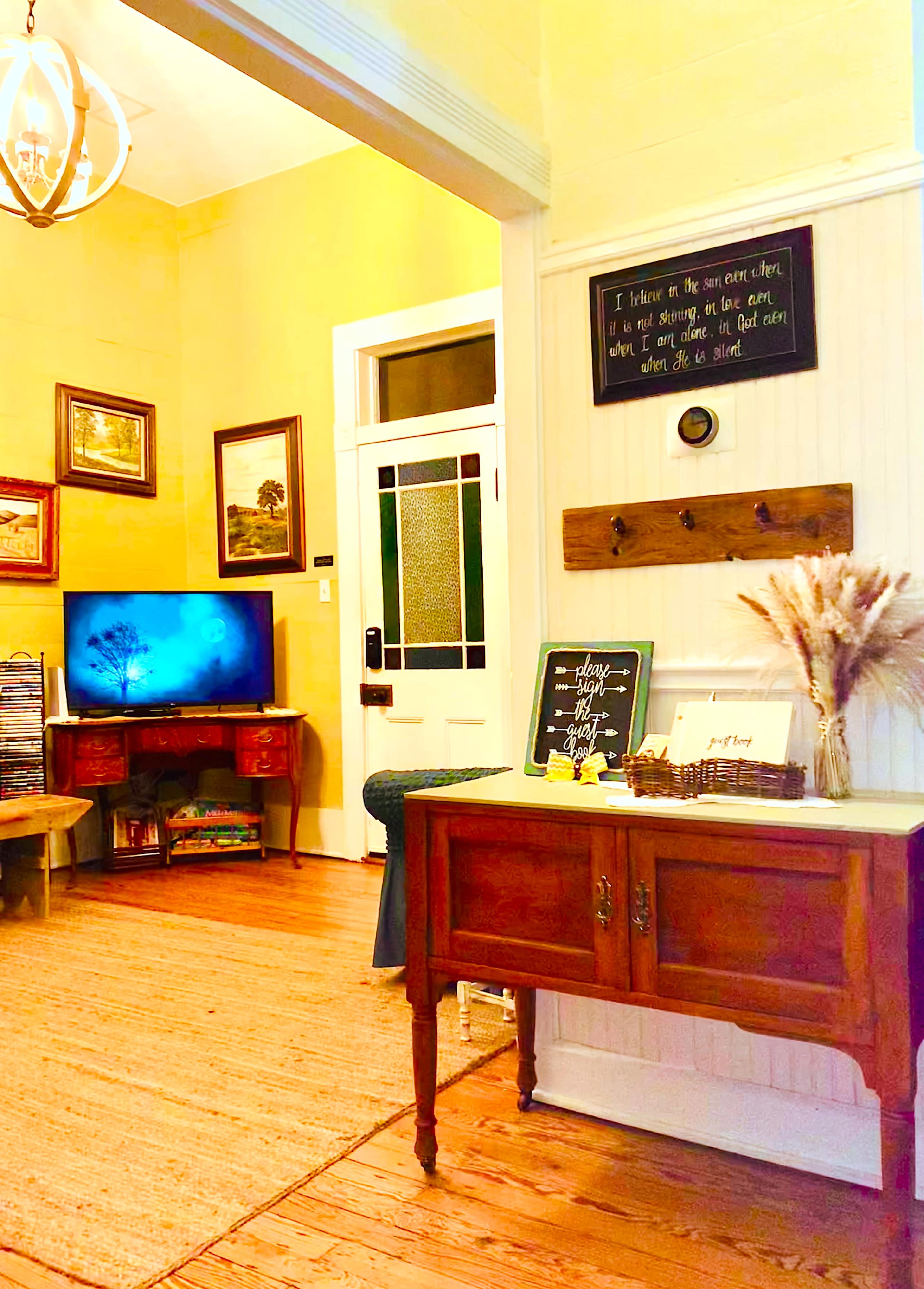 The image shows a cozy living room area featuring a vintage wooden cabinet, a television displaying a nature scene, and decorated walls with framed art and decorative accents.