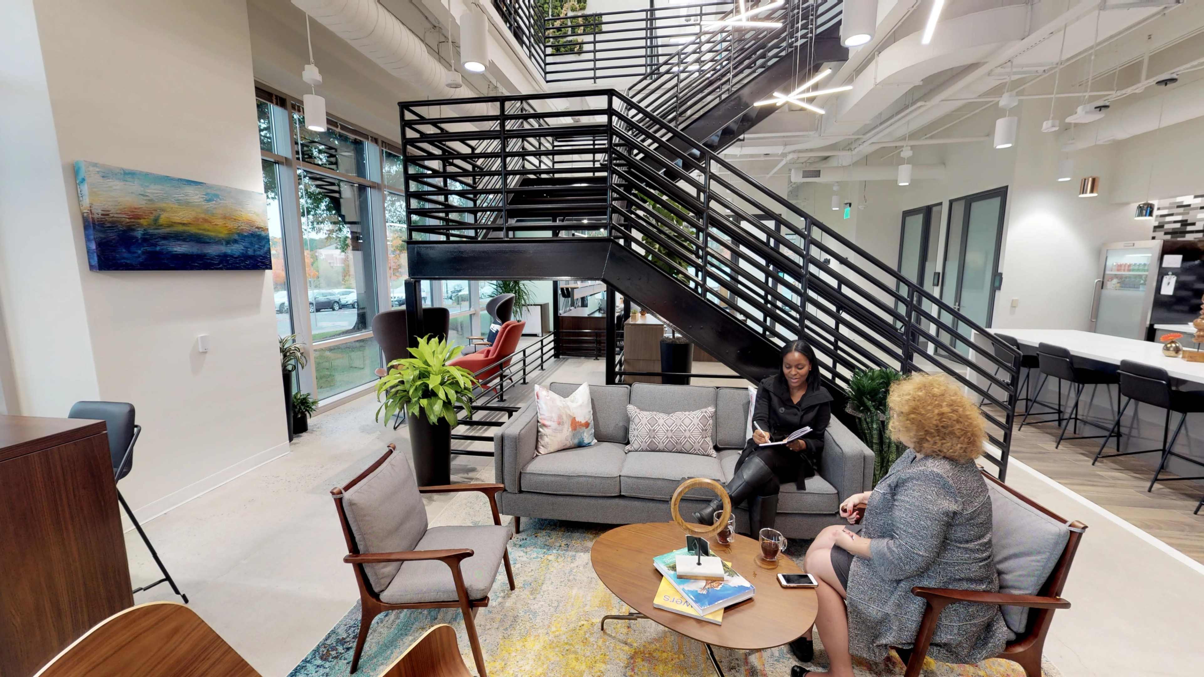 The image shows a modern lounge area with a gray sofa, two chairs, and a round coffee table, accompanied by a staircase and large windows allowing natural light to enter the space.