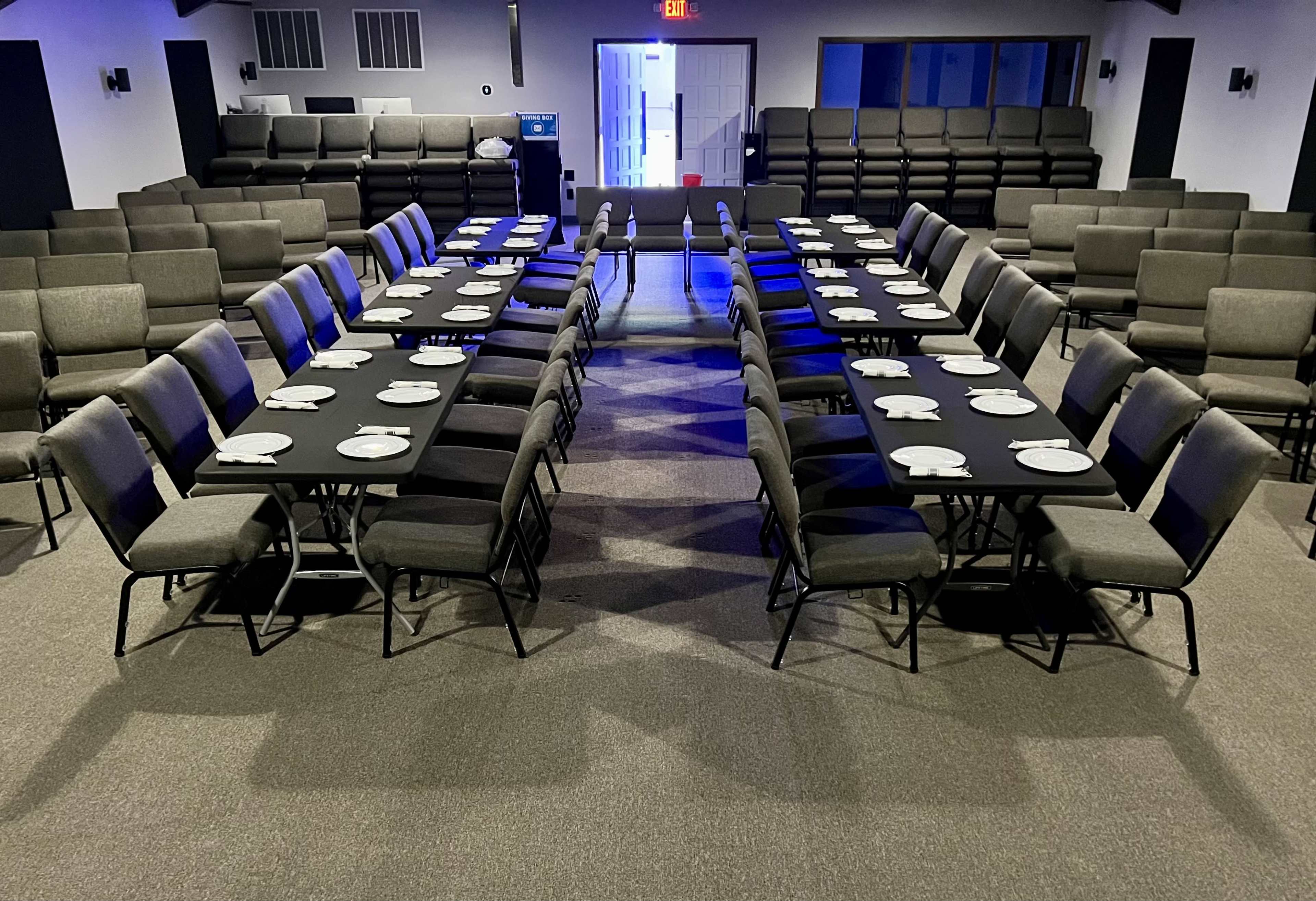 The image shows a dining setup with several long tables arranged in two rows, each covered with black tablecloths and set with plates, in a large, well-lit room with chairs stacked along the walls.